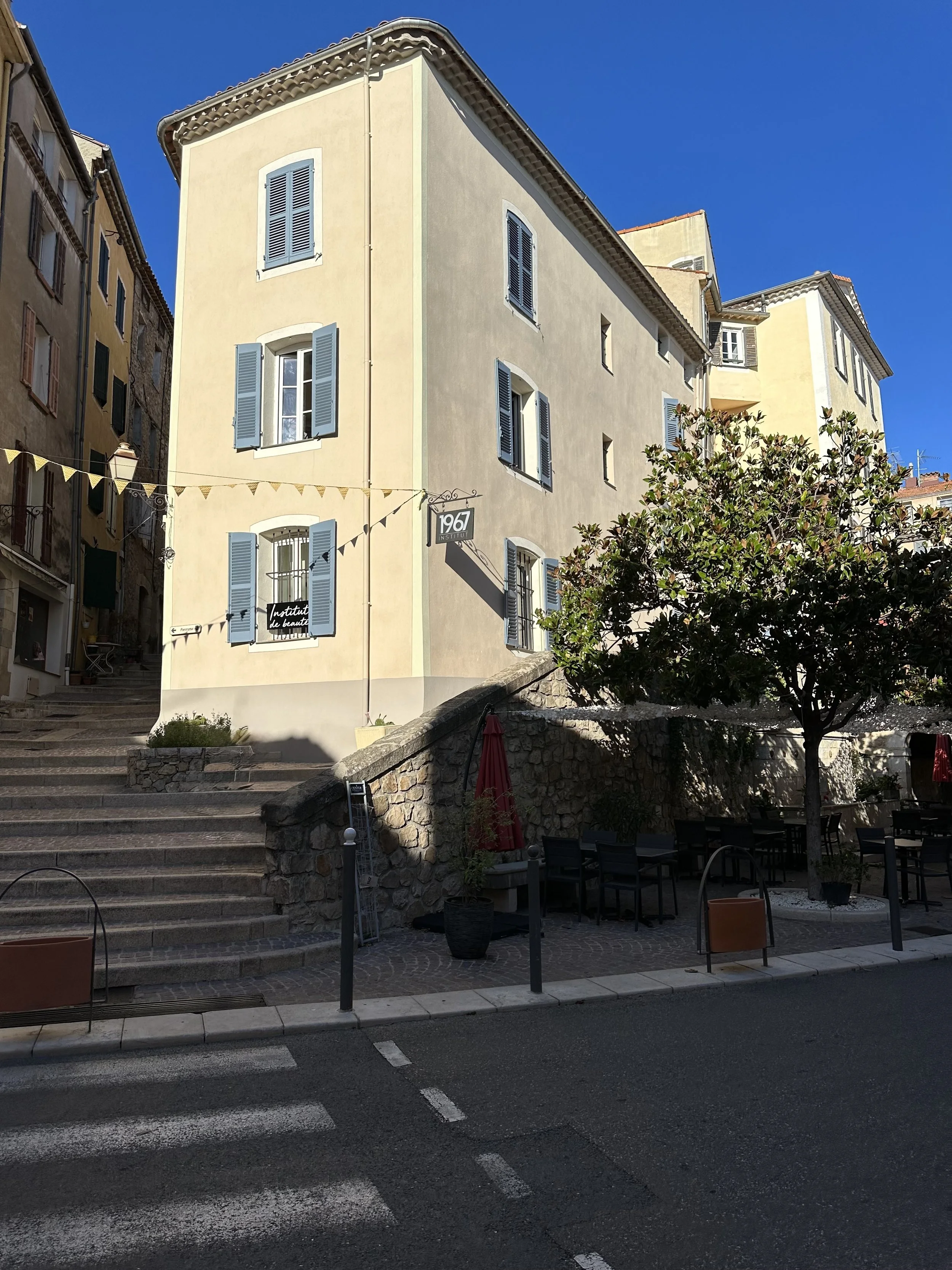 Une rue piétonne avec des escaliers en pierre menant à un bâtiment beige avec des fenêtres à volets bleus, un arbre et des tables avec des chaises en plein air, sous un ciel bleu clair.