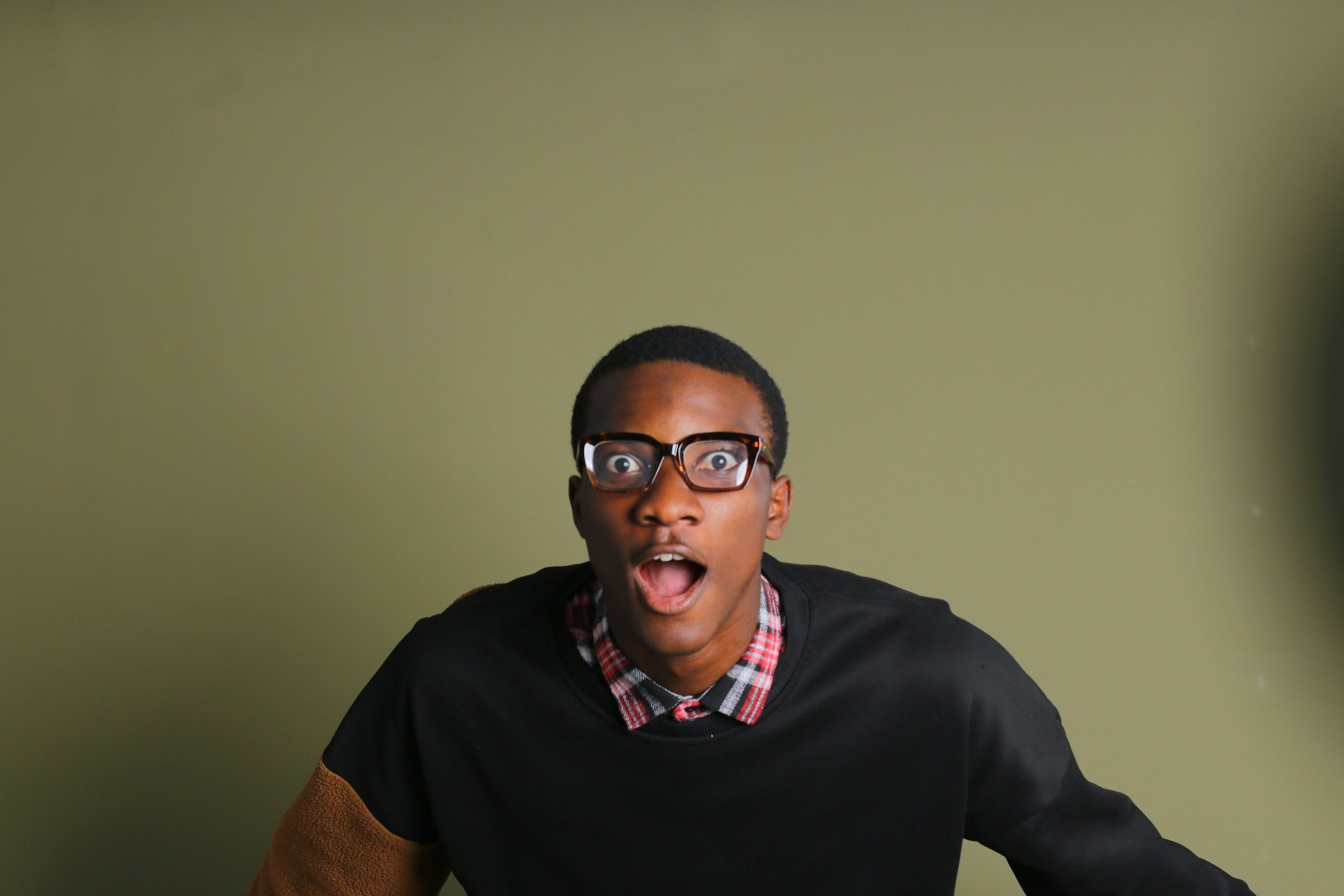 A young man with glasses and short hair, wearing a plaid shirt under a black sweater, shows an expression of surprise or shock with his mouth open and eyes wide, against a plain green background.