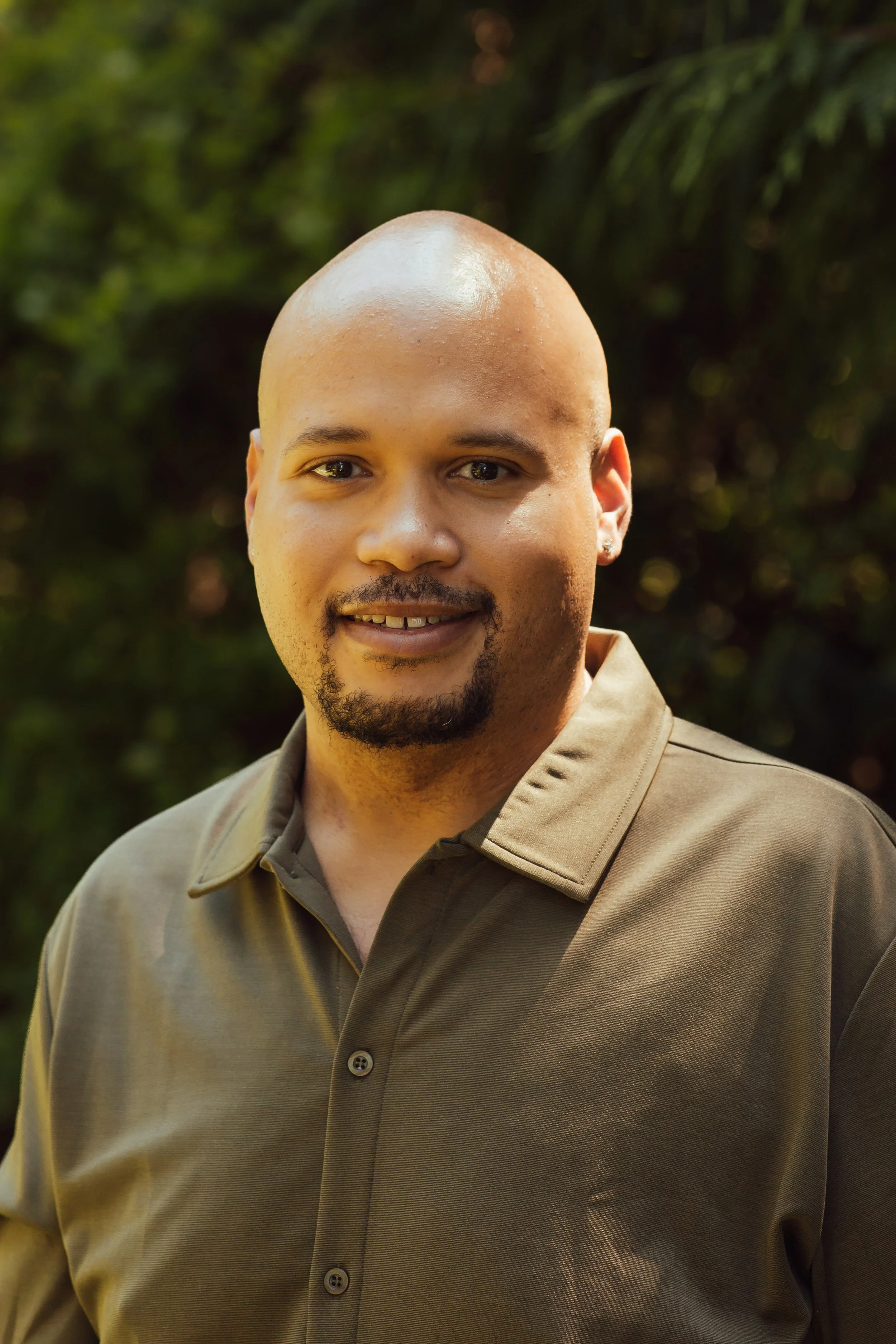 A man with a shaved head, a goatee, and a mustache, smiling outdoors in front of greenery, wearing a brown button-up shirt.