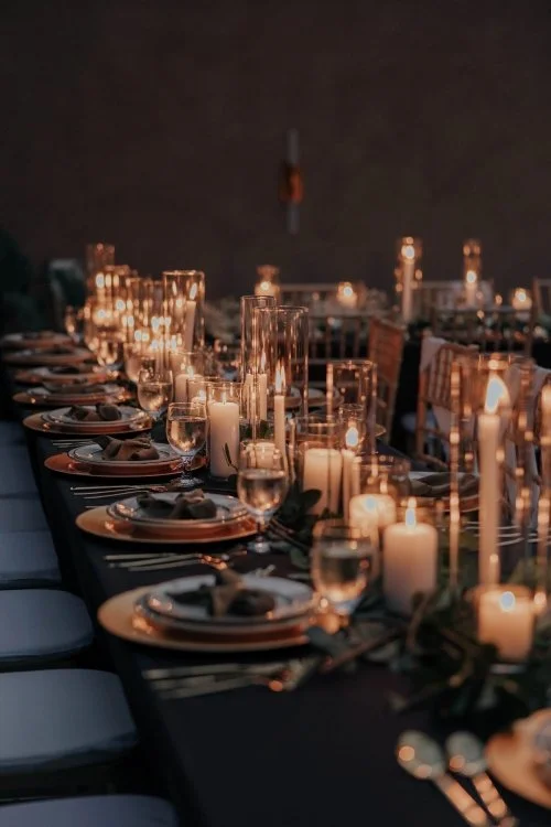 Elegant dining table set for a wedding reception with gold Chiavari chairs, black tablecloth, white plates, gold chargers, green bow napkins, glassware, and numerous candles creating a warm ambiance.