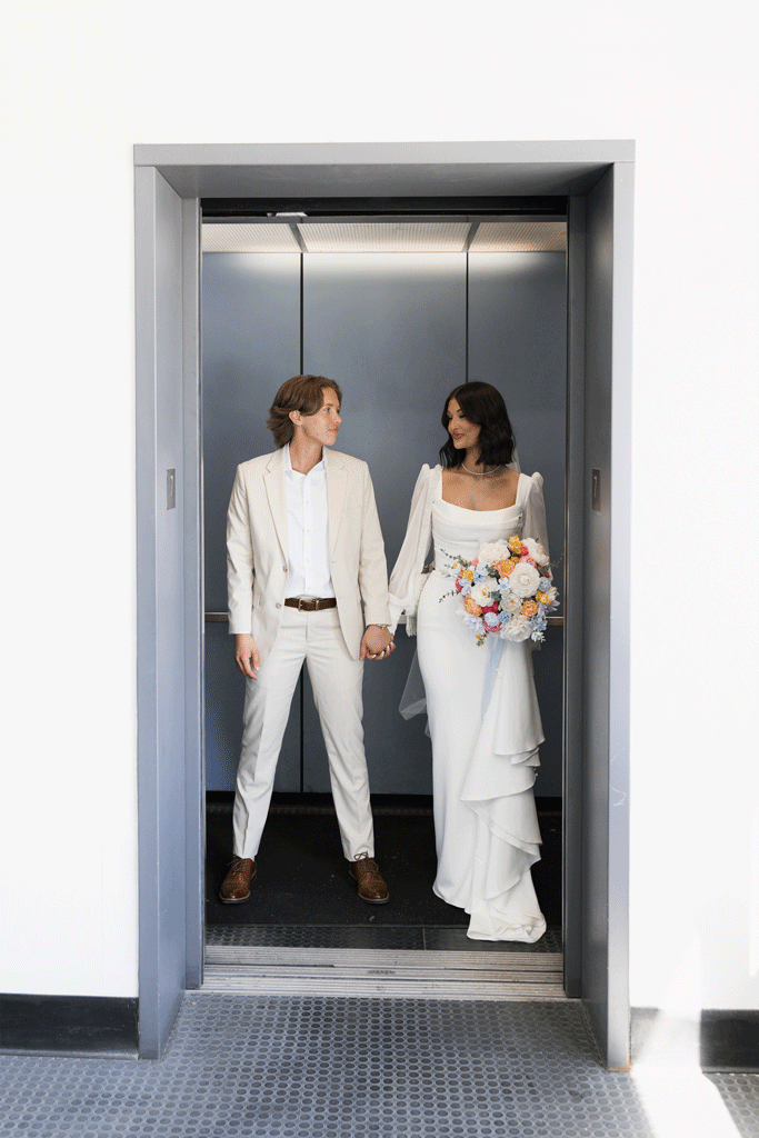 Two women holding hands in an elevator, one in a white suit and the other in a wedding dress holding a bouquet of flowers.
