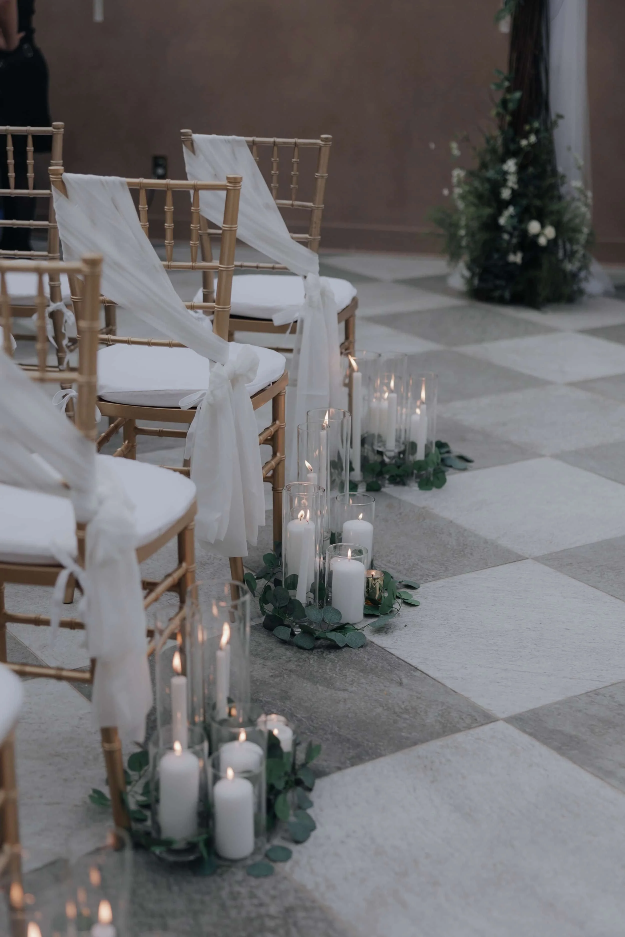 Chairs decorated with white fabric ribbons, set in rows for a wedding ceremony, with white candles and greenery on the floor.