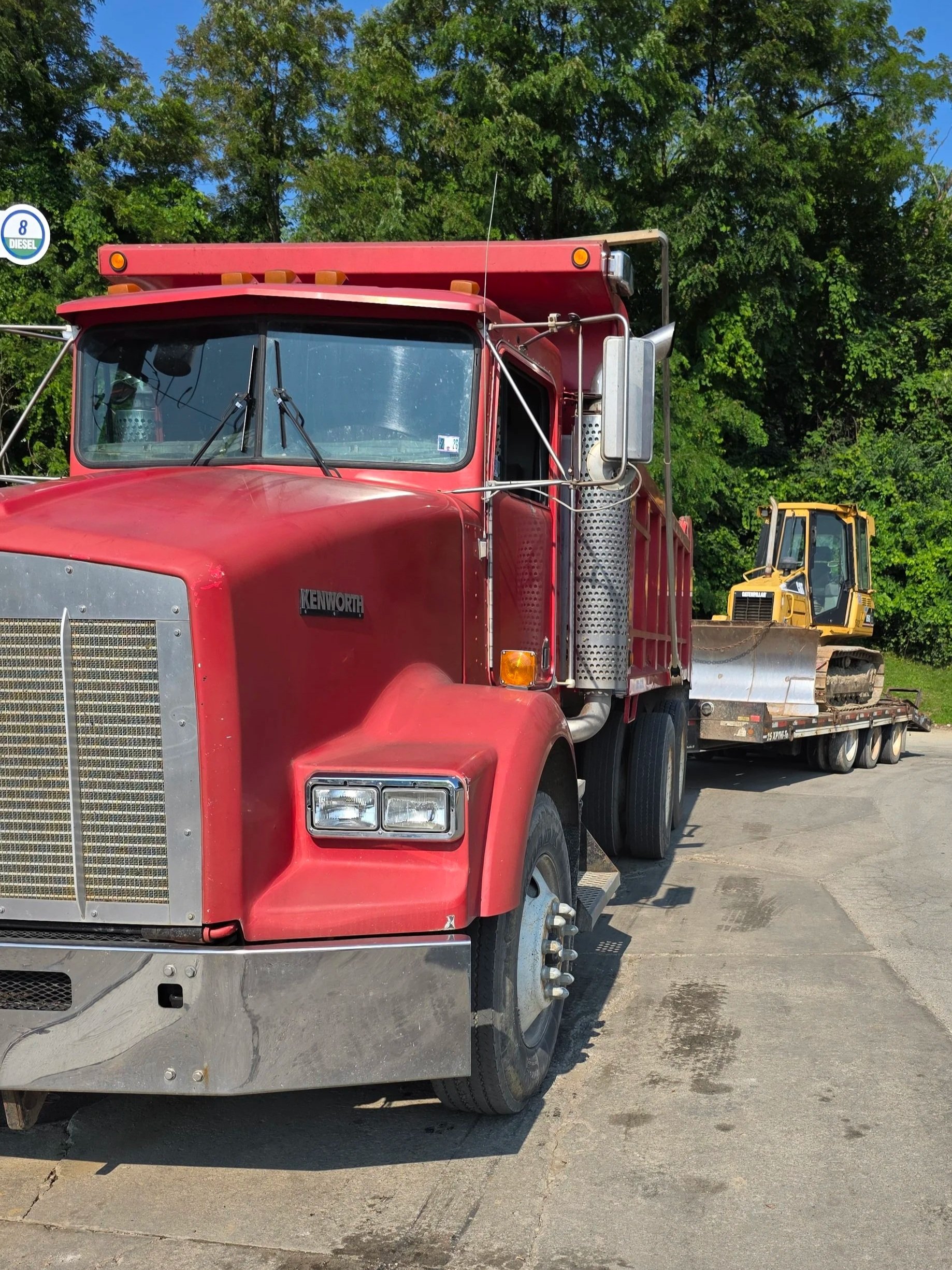 A red Kenworth semi-truck carrying construction equipment, including a yellow bulldozer, parked on a paved surface with green trees in the background.