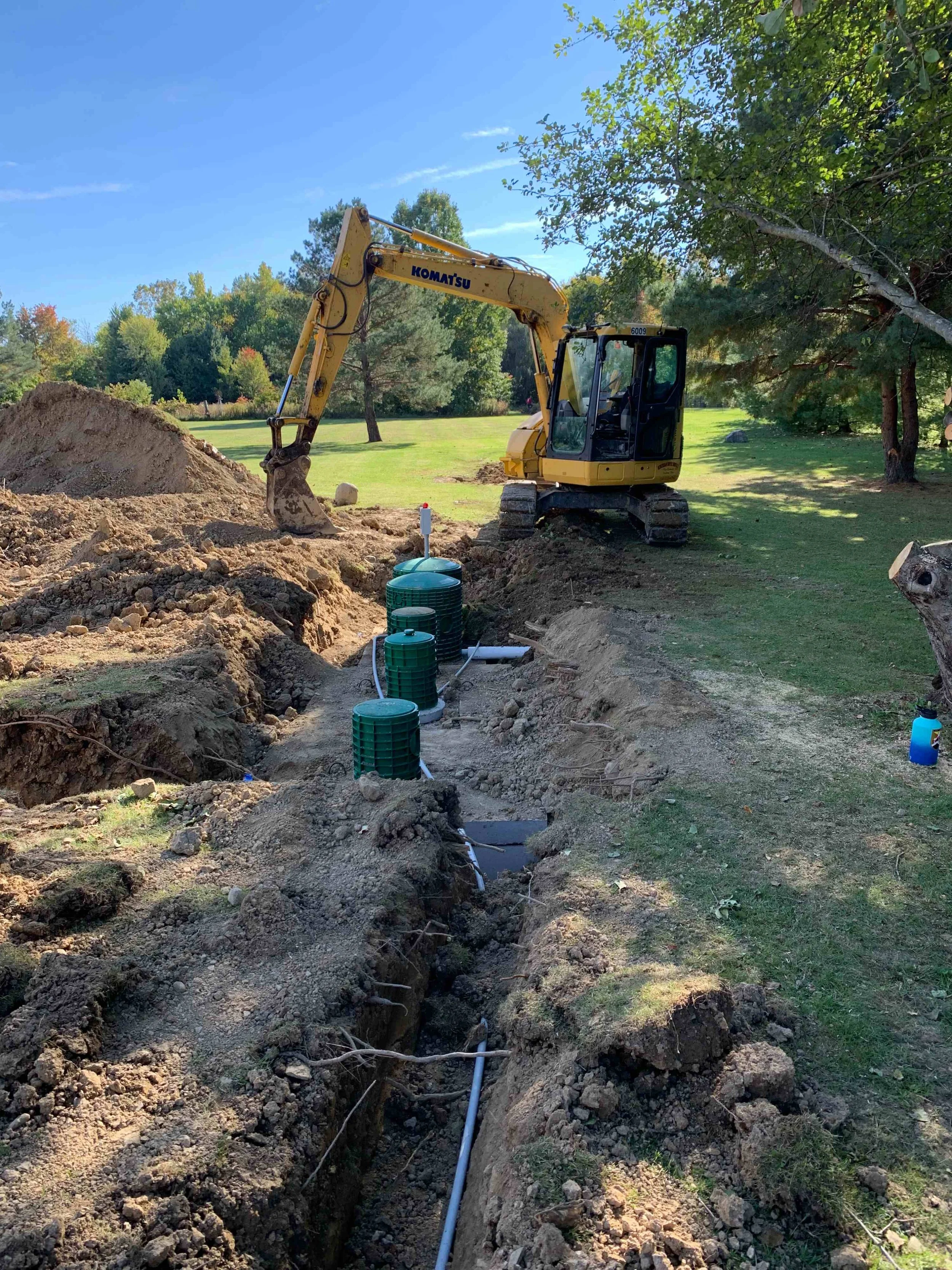 Construction site with a small yellow excavator digging a trench in a grassy area with trees, green water tanks, and pipes.