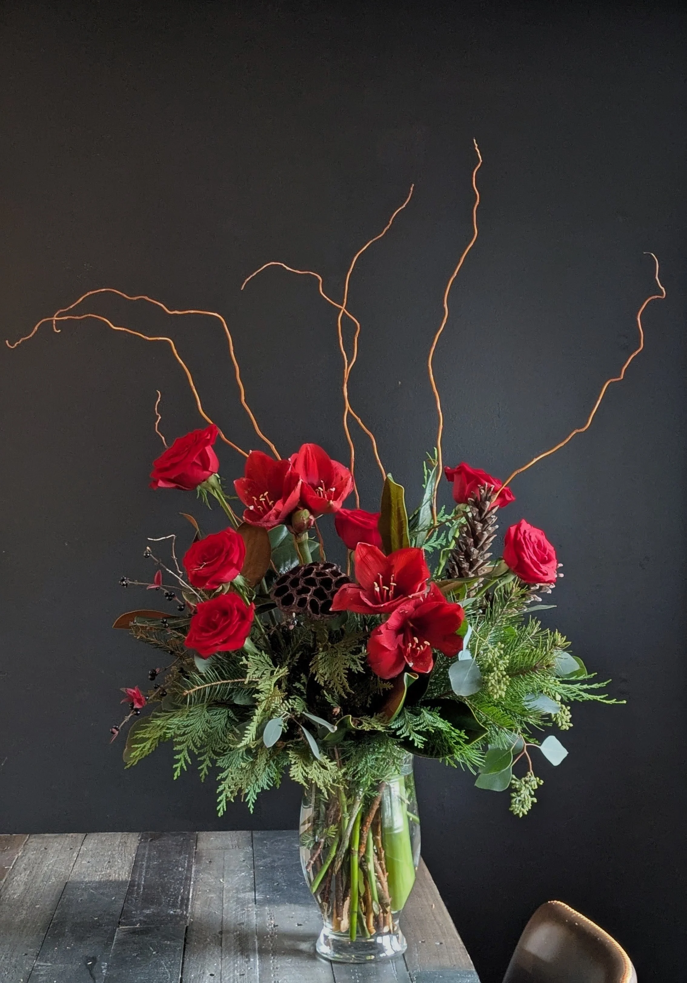 A Holiday floral arrangement with red roses, white calla lilies, white ranunculus, and greenery, placed on a wooden stump in front of a plain beige wall.