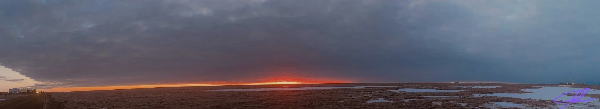 Sunset over a flat marshland with dark clouds in the sky and water patches on the ground.