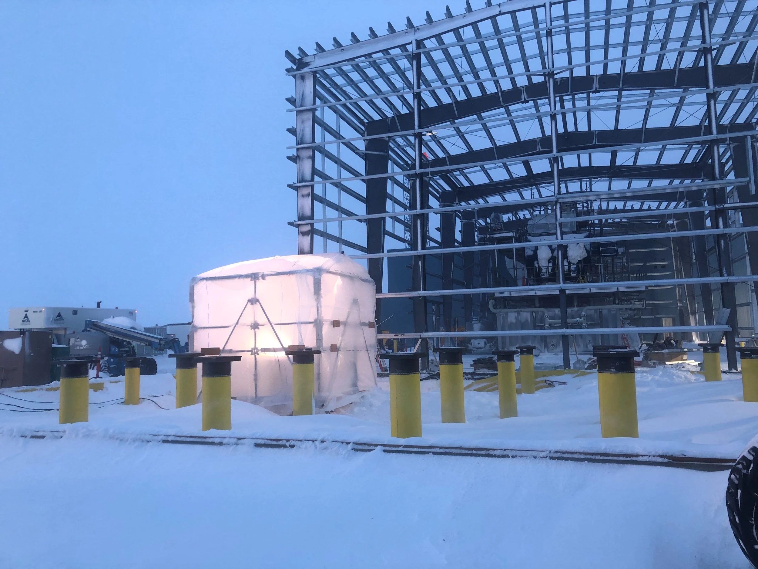 Construction site of a building with a steel frame structure, surrounded by snow, with a partially covered object in the foreground.