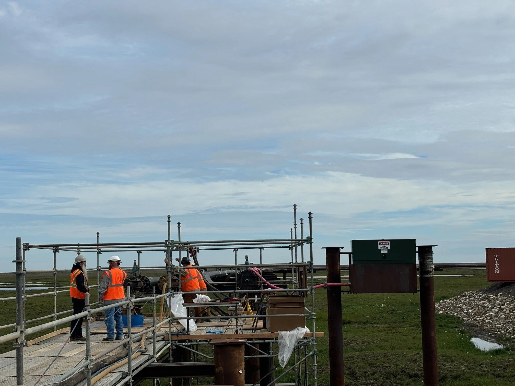 Workers in safety vests and helmets working on construction site with scaffolding and equipment in an open field under cloudy sky.