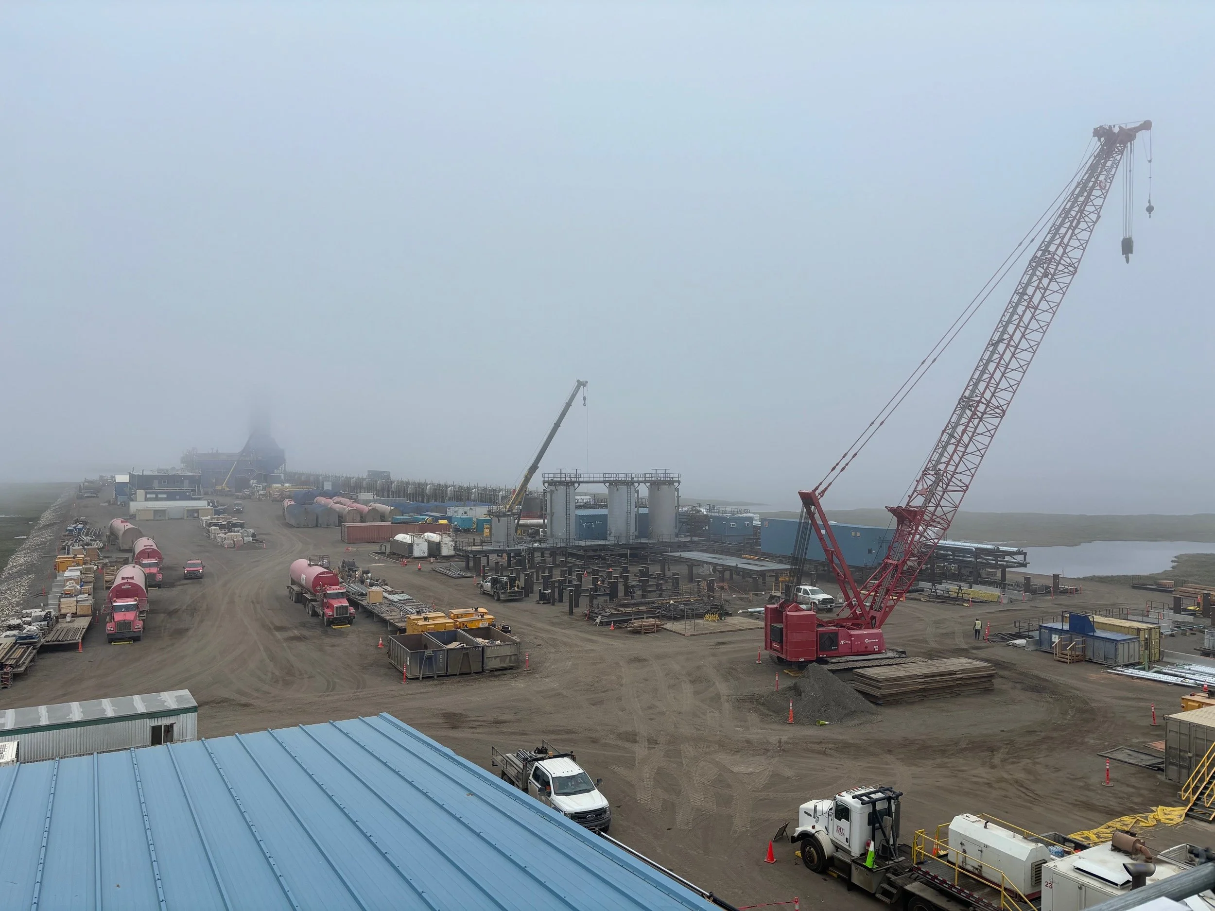 Construction site with cranes, trucks, and equipment in foggy weather near water.