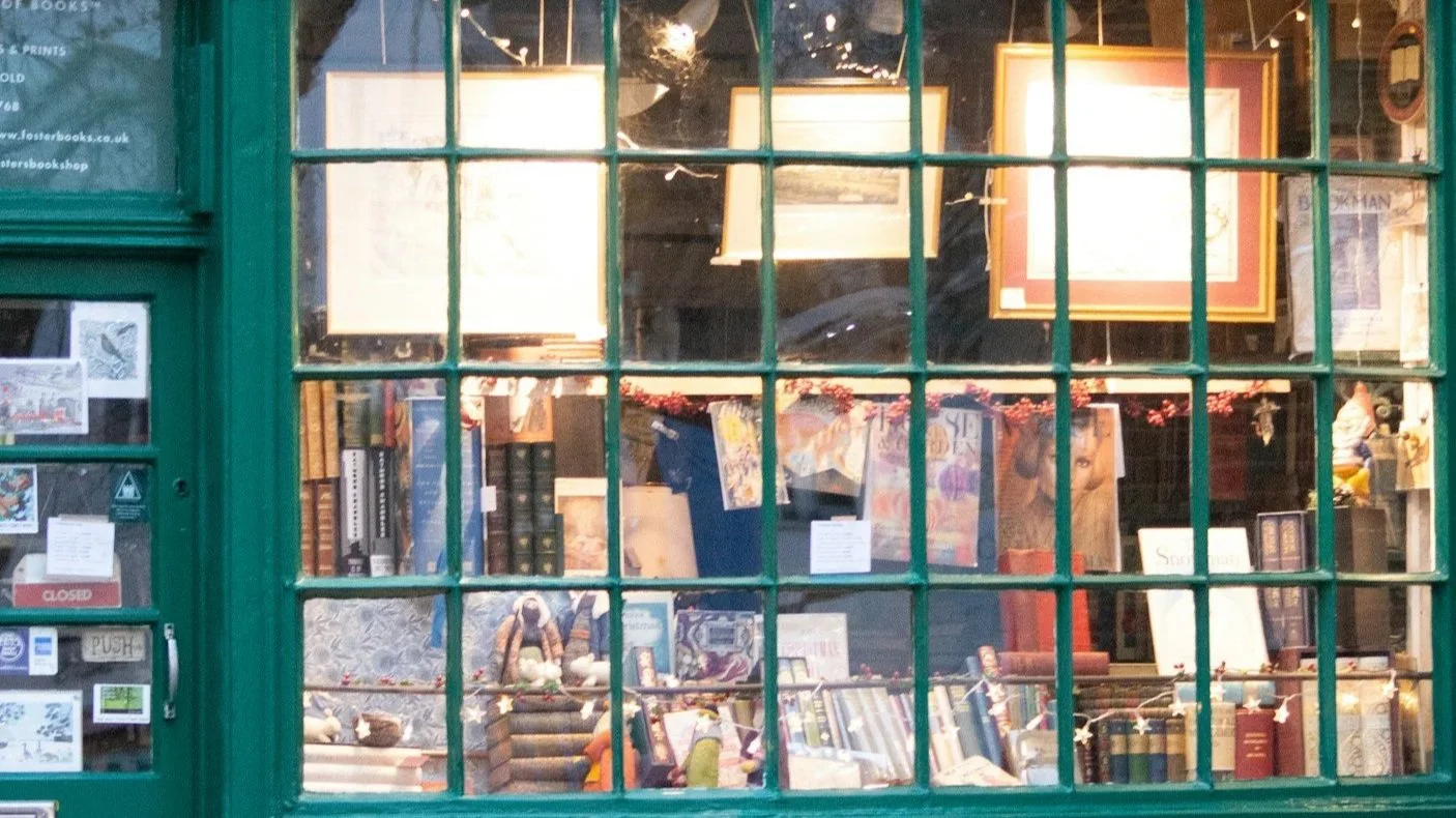View of a bookstore through a green window with multiple panes, displaying books, framed pictures, and decorative items inside.