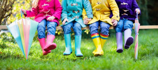 Four children sitting on a bench wearing colorful raincoats and rain boots, holding umbrellas in a park.
