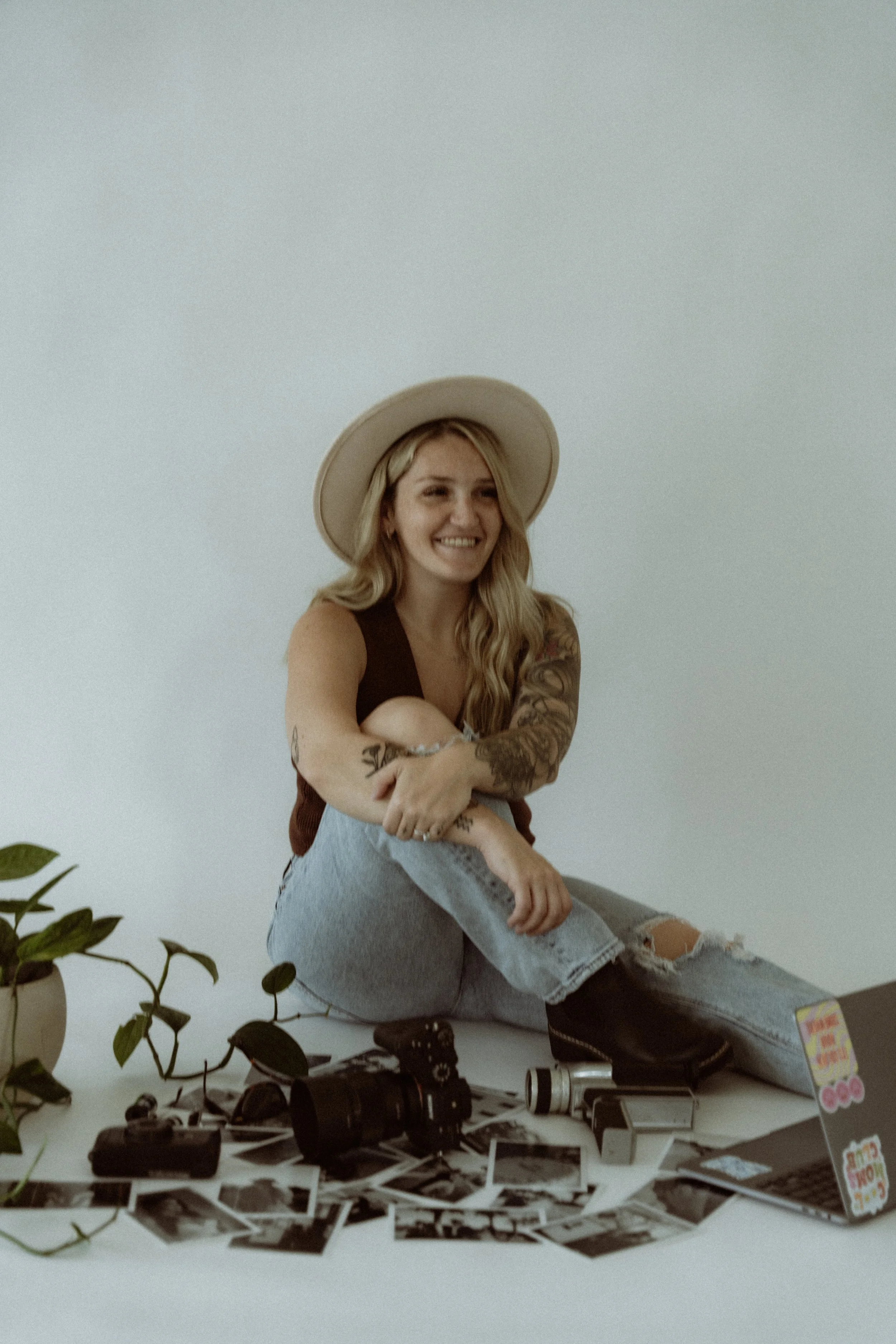 A young woman with long blonde hair, tattoos, wearing a hat, sitting on the floor surrounded by photos, a camera, a laptop with stickers, a potted plant, and a flashlight, smiling at the camera.