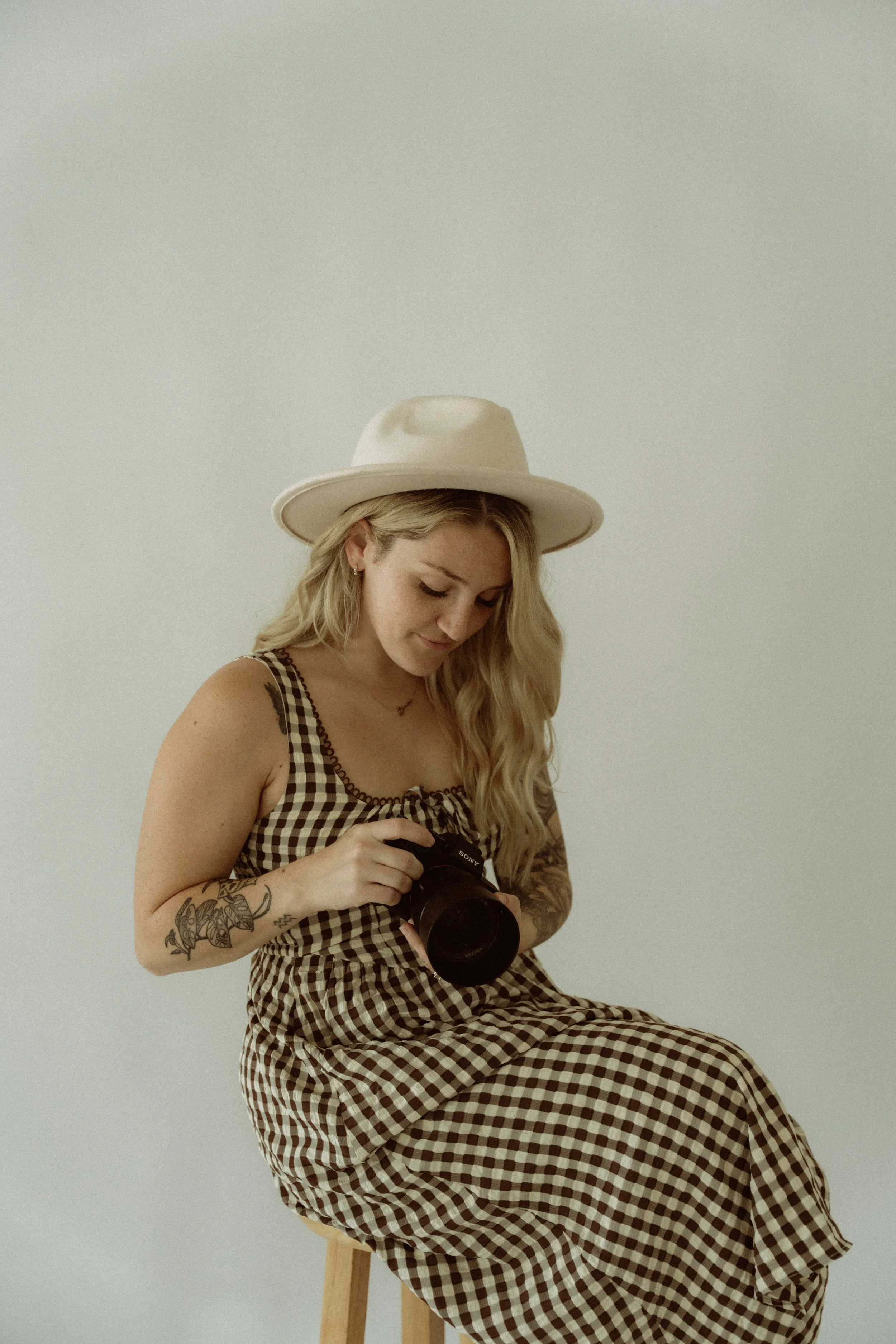 A woman with long blonde hair and tattoos on her arm, wearing a checkered dress and a wide-brimmed hat, sitting on a stool and looking at a camera.