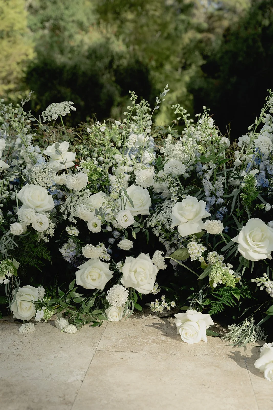 White floral arrangement with roses and various white flowers on a stone surface, with a blurred green natural background.