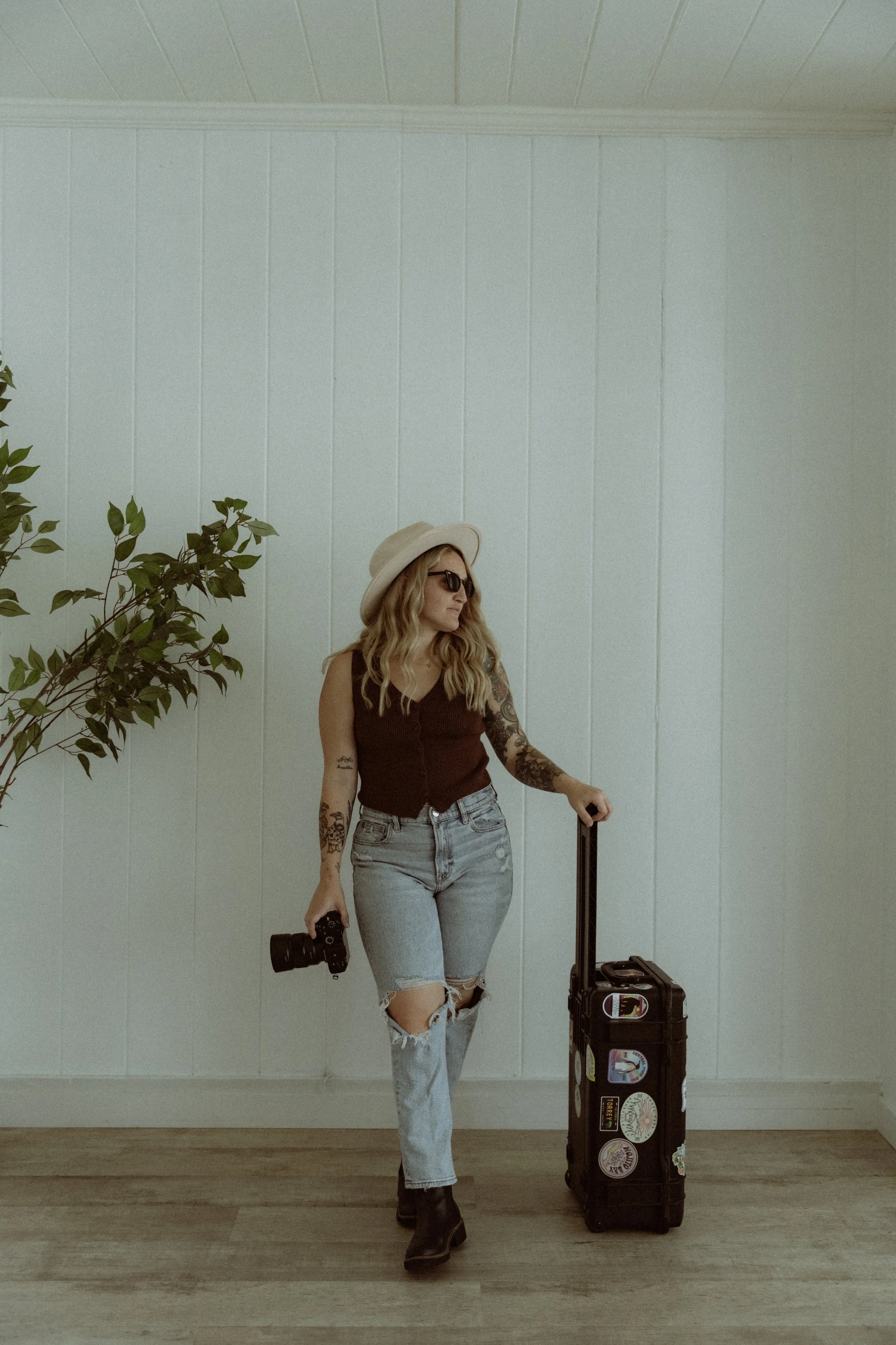 A woman with blonde wavy hair, wearing a beige hat, sunglasses, a sleeveless dark top, ripped jeans, black boots, and tattoos on her arms, stands indoors against a white paneled wall. She is holding a black camera in her right hand and has a black rolling suitcase decorated with colorful stickers.