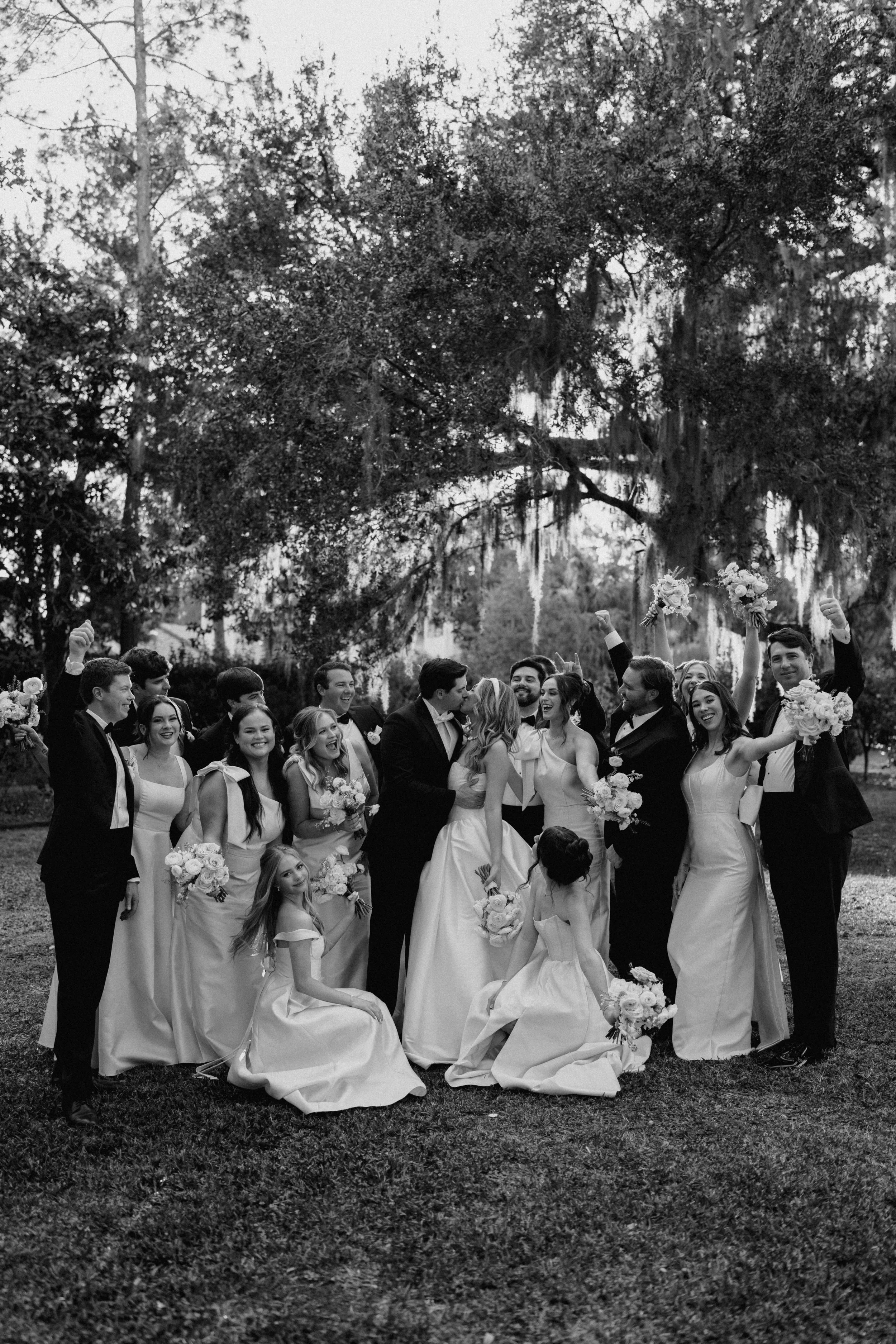 A group of people in formal attire celebrating at a wedding outdoors under large trees, with some holding bouquets and raising their hands in celebration.