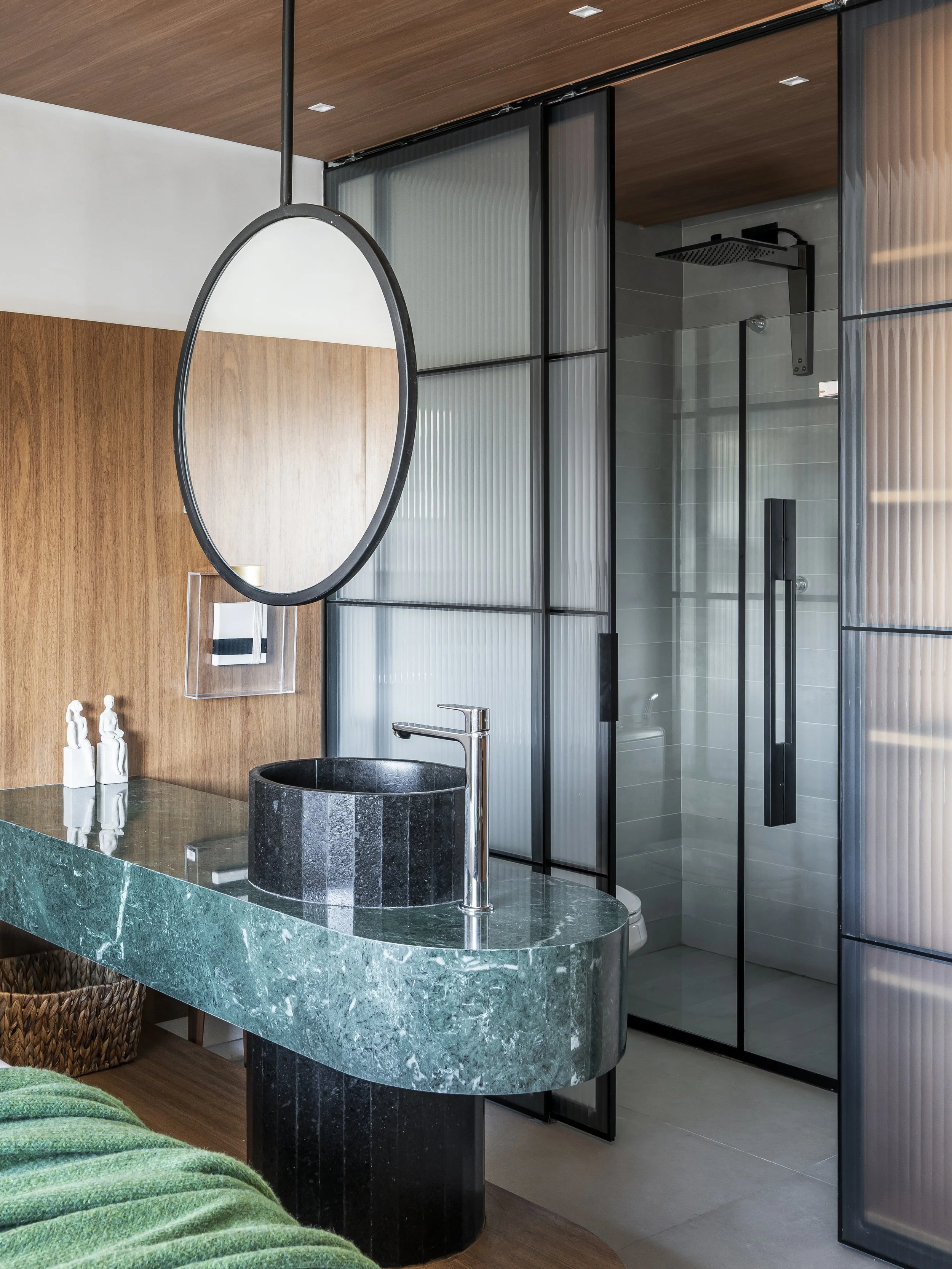 Banheiro moderno com pia de tampo verde, espelho oval, parede de madeira e box de vidro com portas de correr.