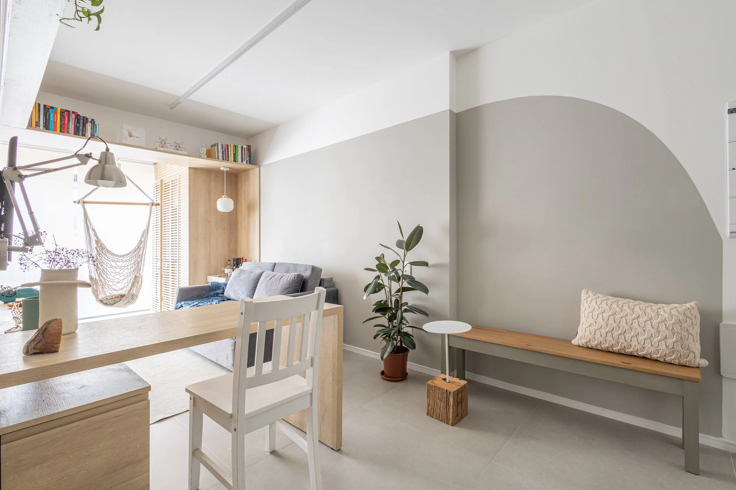 Sala de estar com móveis de madeira clara, sofá cinza, plantas, prateleira com livros, pufe de madeira, acompanhamento de decoração minimalista branca e cinza.