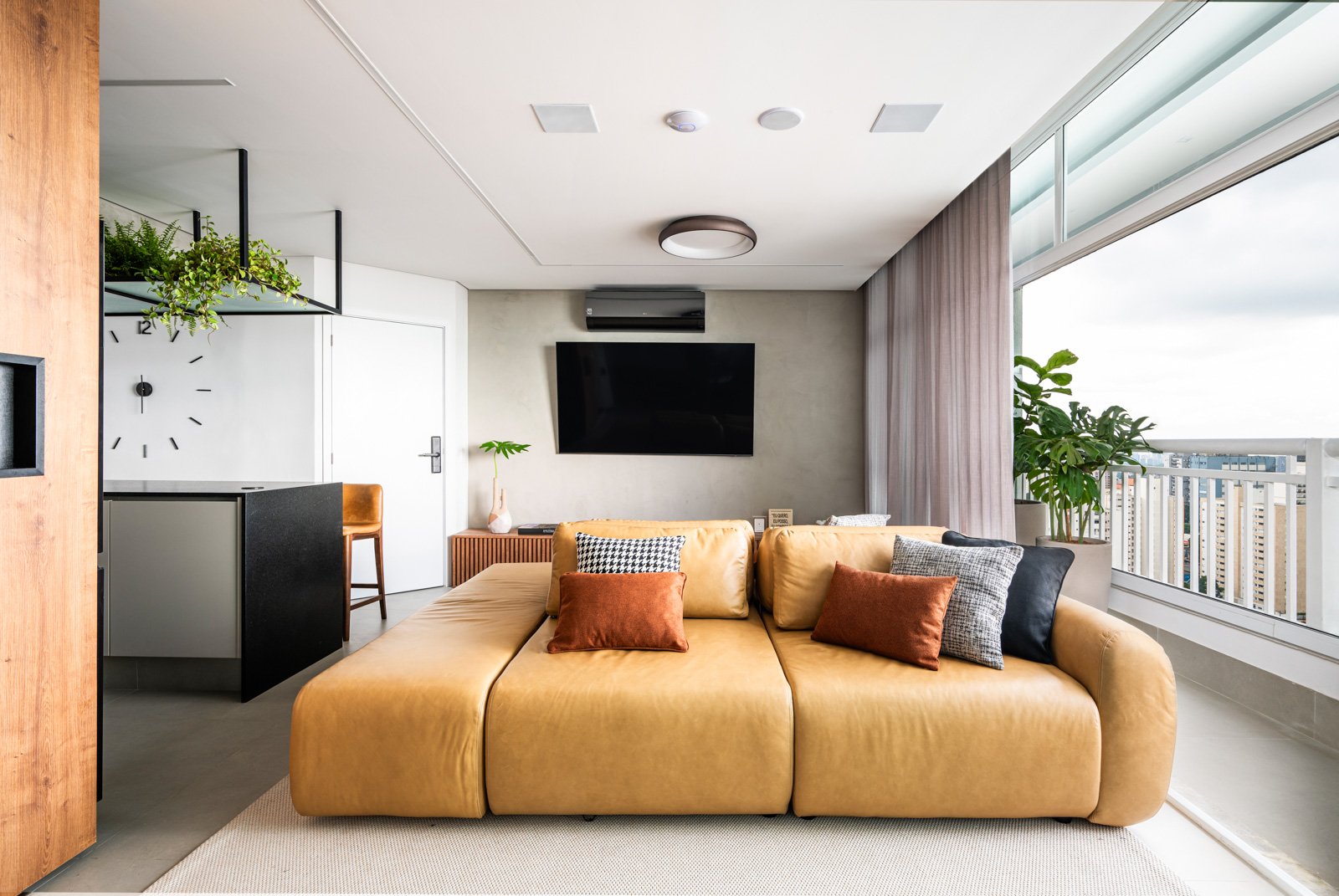 Salão de apartamento com sofá amarelo, janelas grandes com vista para prédios, televisão na parede, plantas, cortinas e decoração moderna.
