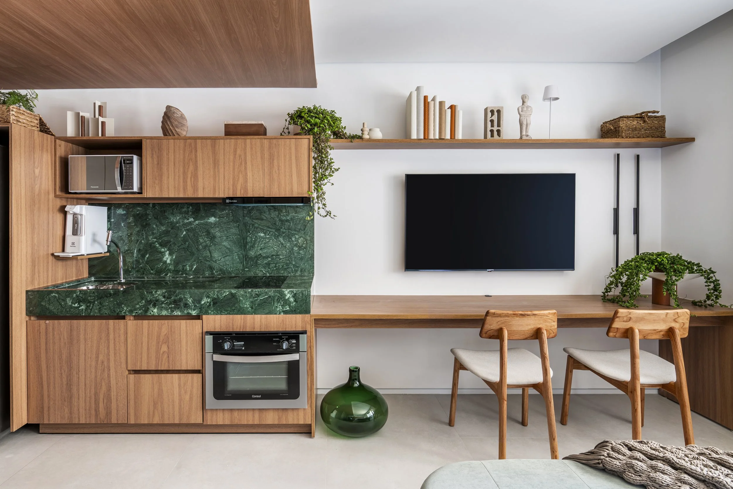 Sala de estar com cozinha integrada, móveis de madeira, televisão de tela plana na parede, bancada de madeira, duas cadeiras de madeira com assento claro, gaveta com forno embutido, micro-ondas, armário com pia de mármore verde escuro, prateleira com livros, plantas e decoração