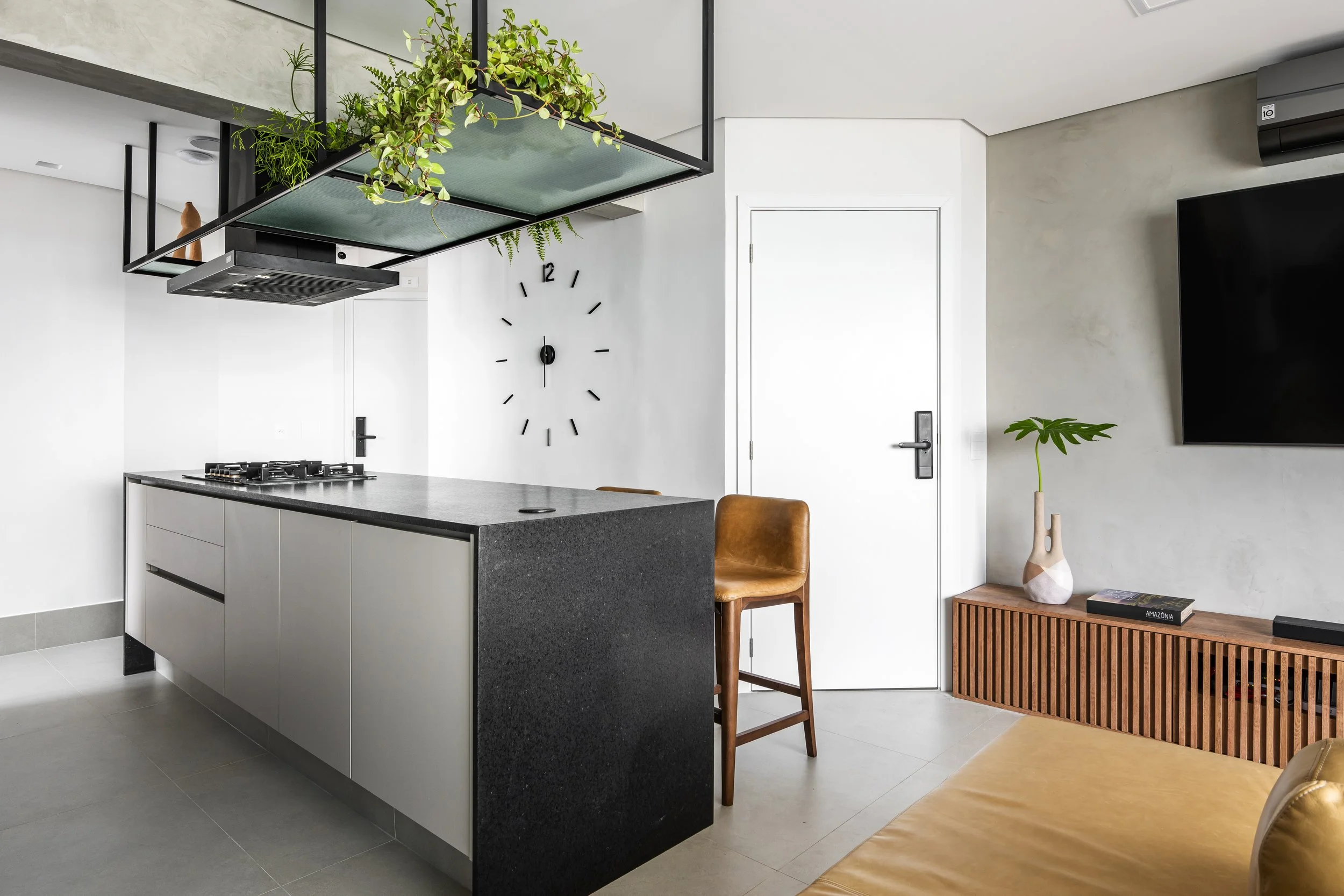 Imagem de uma sala de estar moderna com área de cozinha ao fundo, parede branca, relógio de parede, porta branca, televisão e uma mesinha de madeira com vaso e planta. Existem móveis de cadeira em tom de caramelo e uma poltrona dourada.