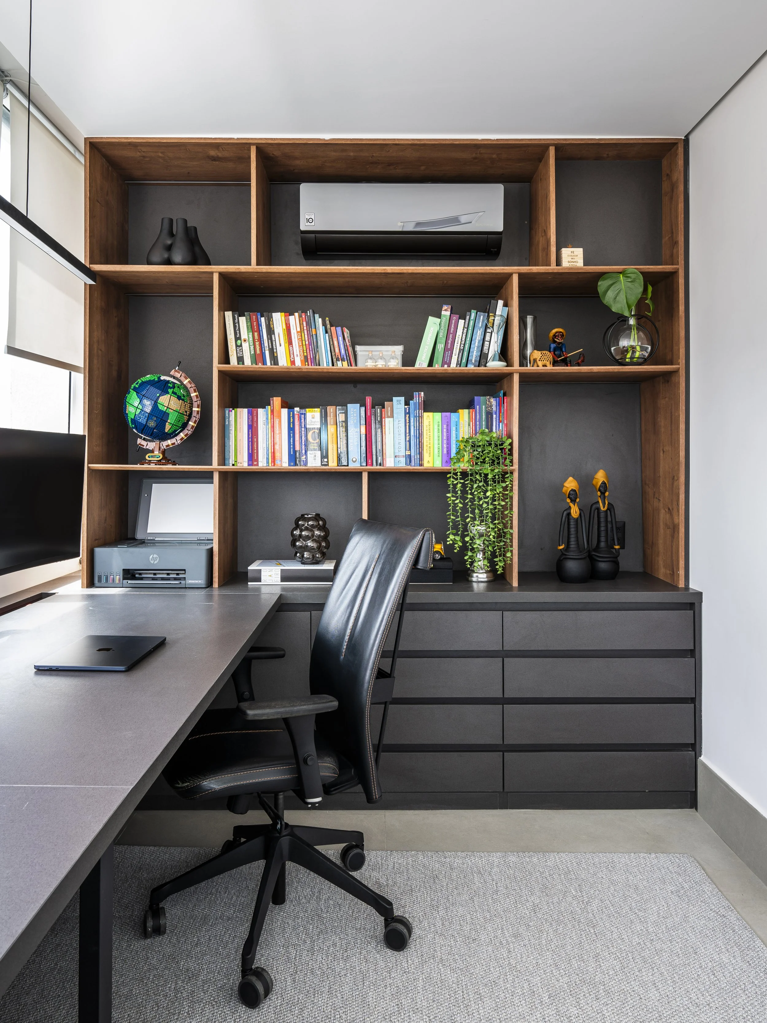 Estante de madeira com livros, objetos decorativos e plantas, em um escritório com cadeira e mesa