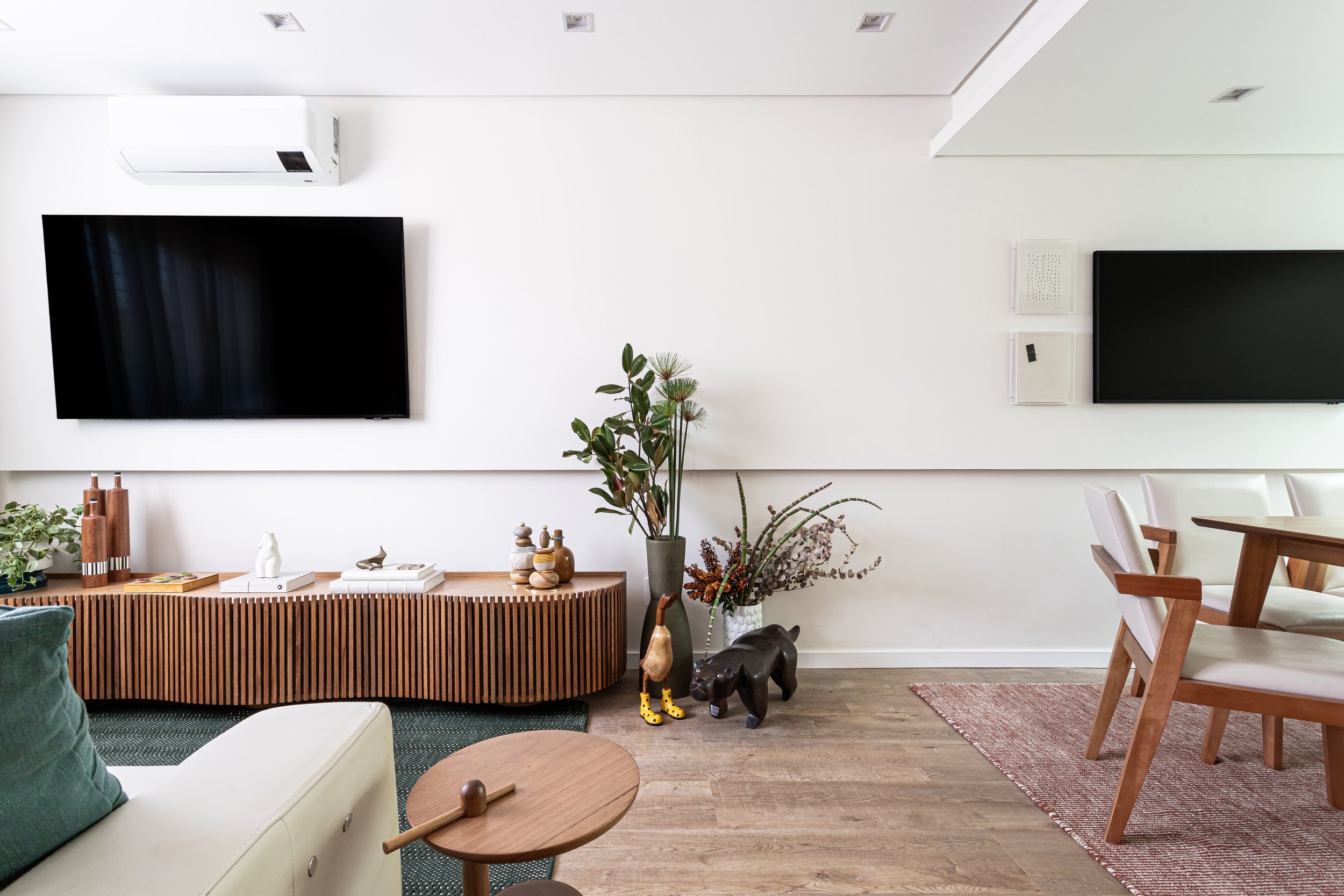 Sala de estar com televisão de tela plana na parede branca, mobiliário de madeira e várias plantas decorativas ao redor.