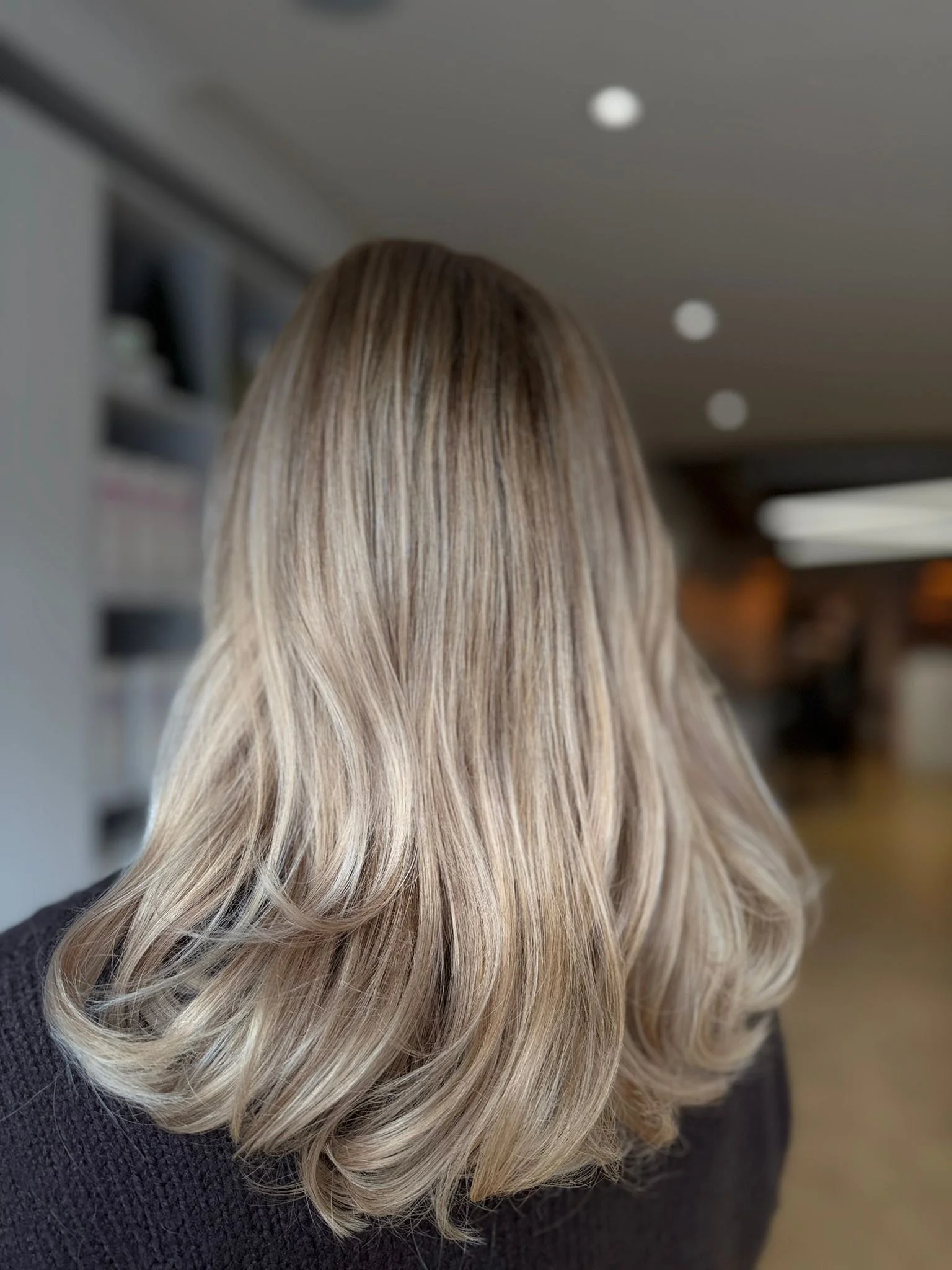 Back view of a woman's long blonde hair with soft curls, indoors with blurred background.