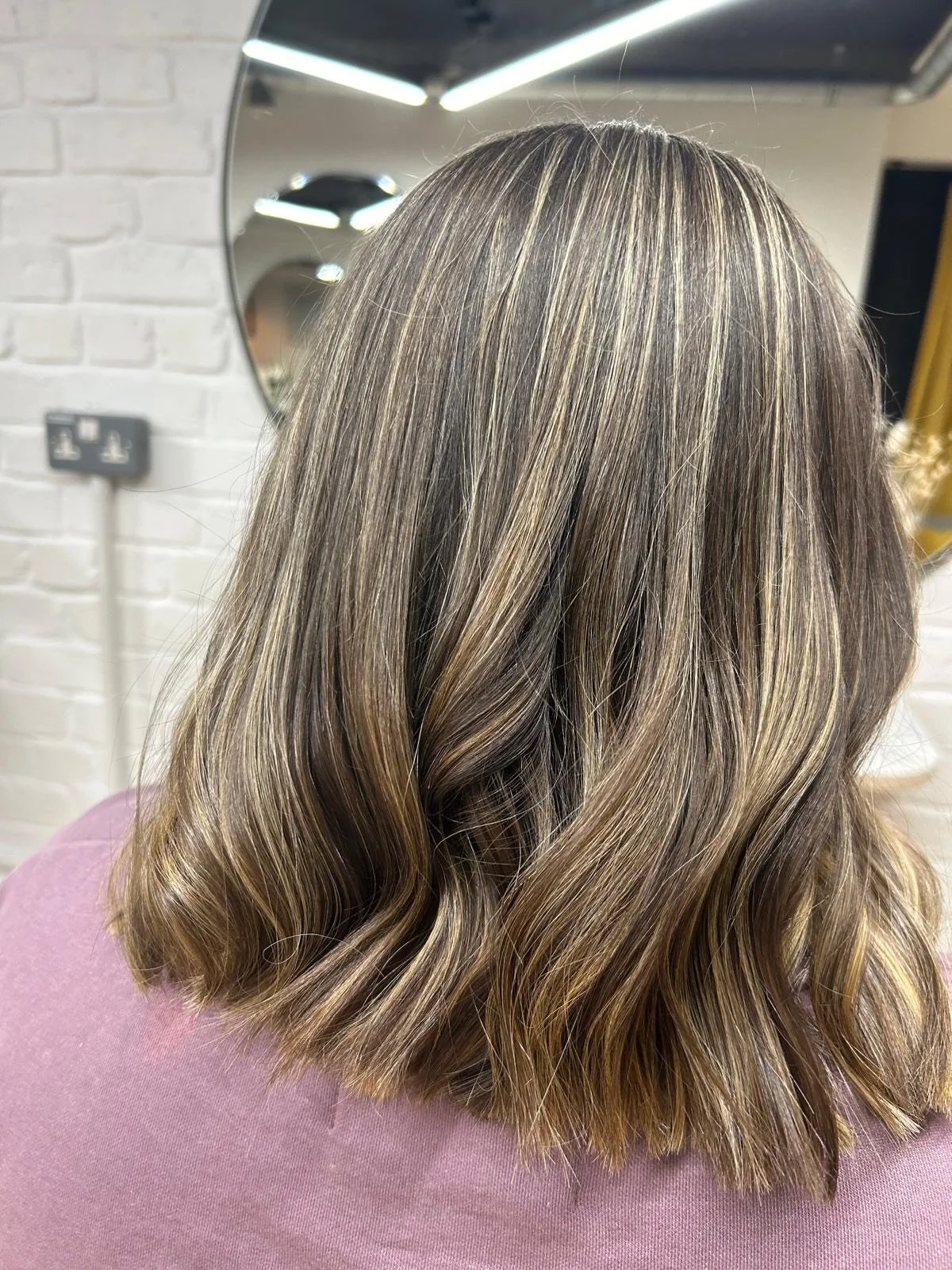 Back view of a woman with shoulder-length, wavy, light brown hair with blonde highlights, wearing a mauve top, in a salon with a mirror and white brick wall.