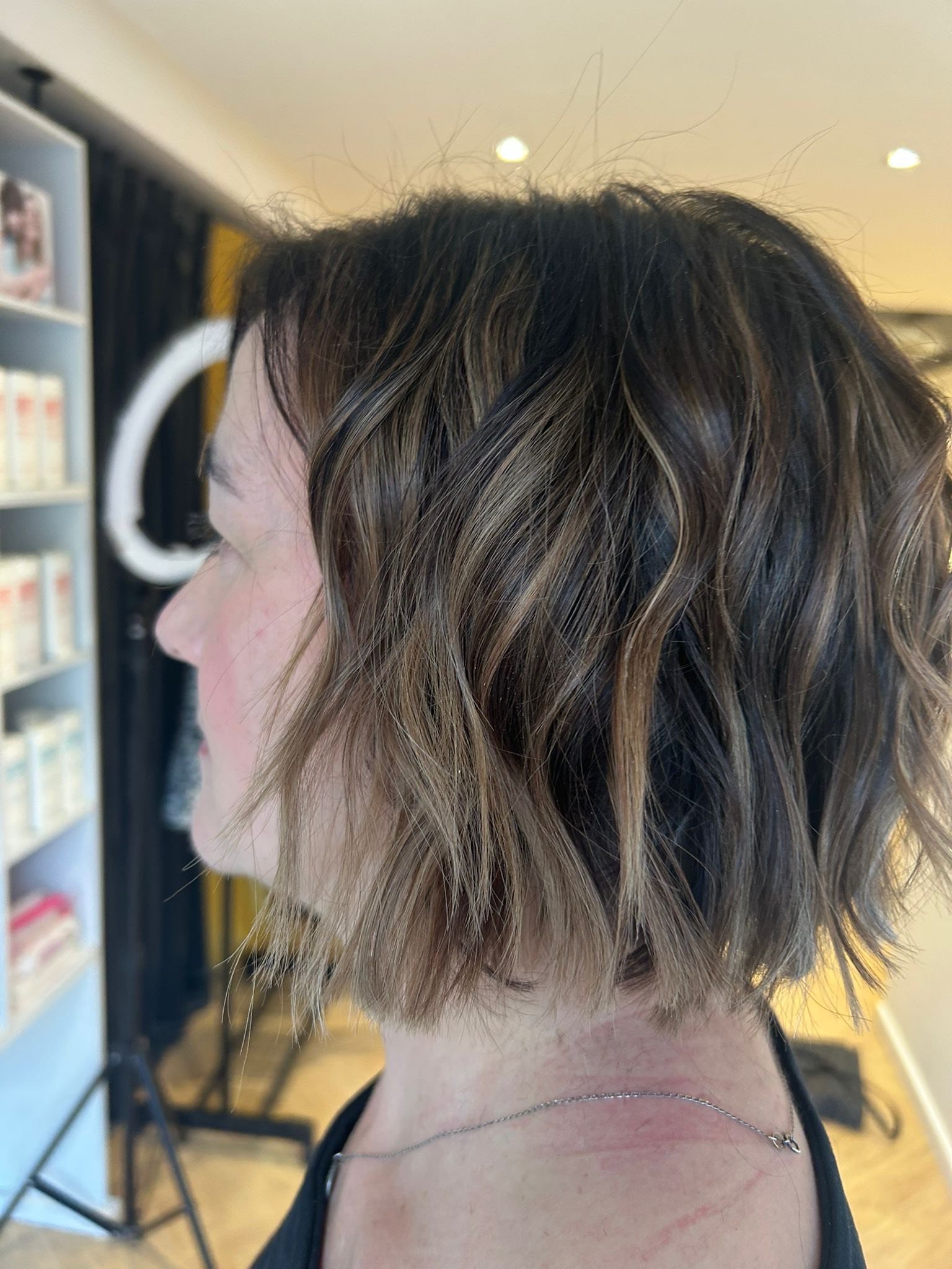 Side view of a woman with short, wavy, brown hair with blonde highlights, wearing a black top and a delicate necklace, in a salon or similar setting.