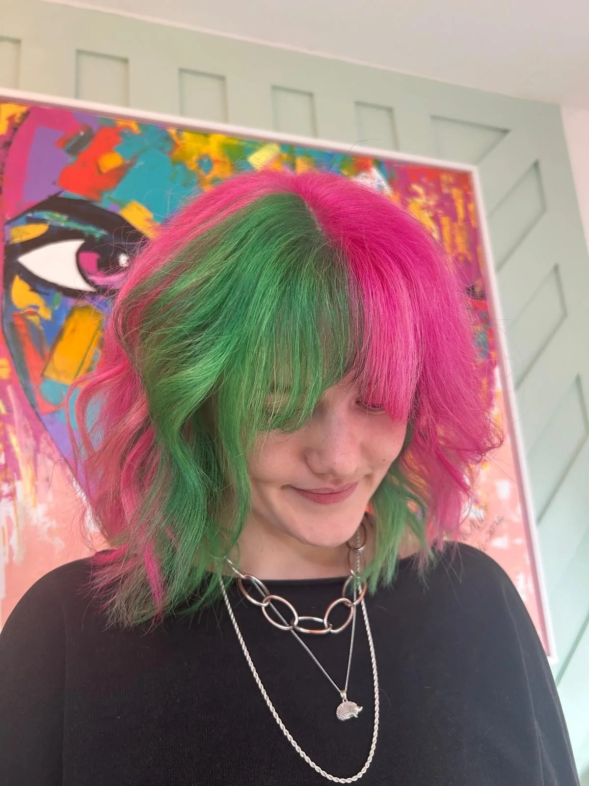 Woman with shoulder-length hair dyed pink and green, smiling with eyes closed, wearing layered silver necklaces and a black top, standing in front of colorful abstract art.
