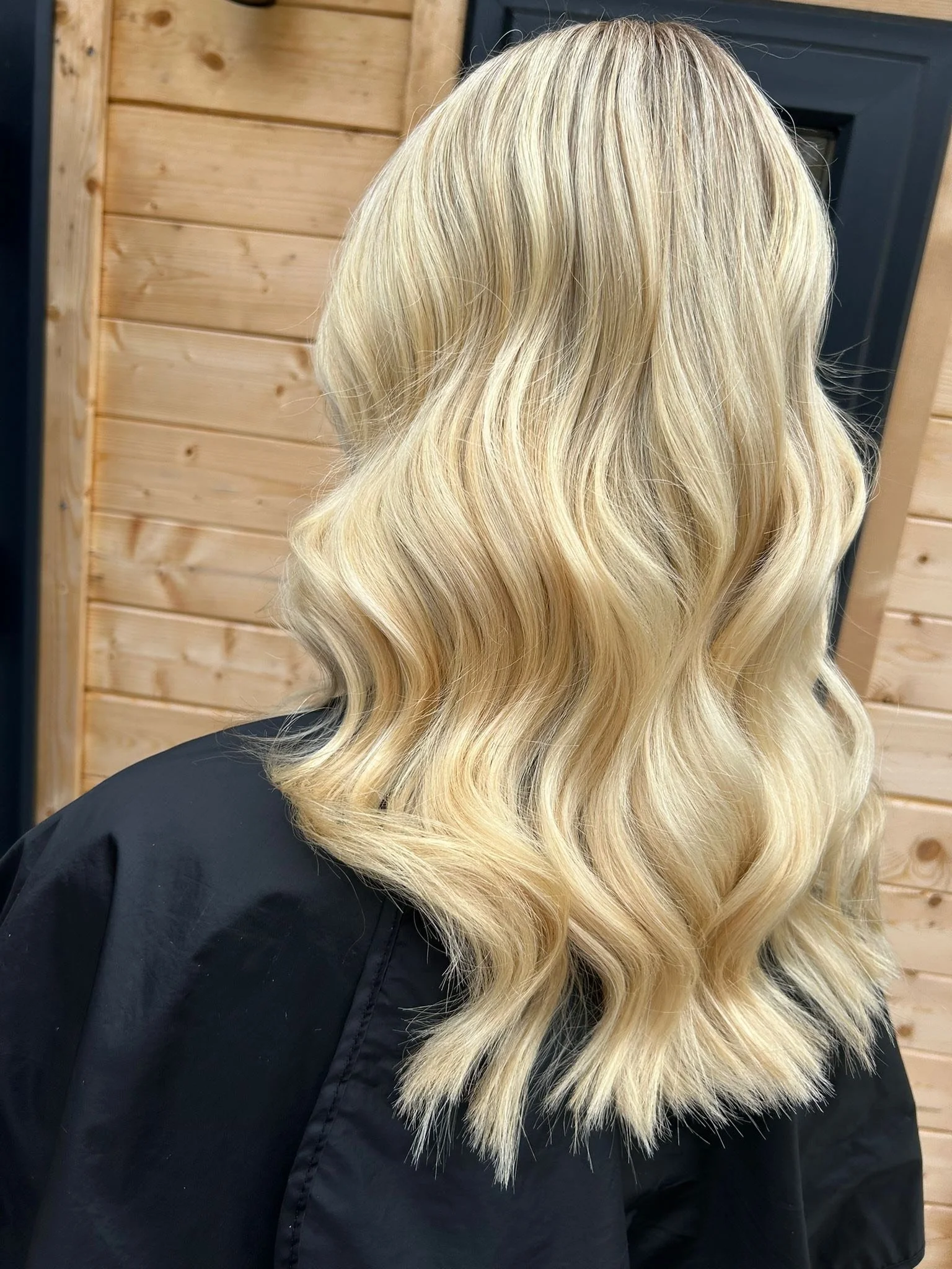 Back of a person with long, wavy, blonde hair, wearing a black cape, standing in front of wooden wall and door.