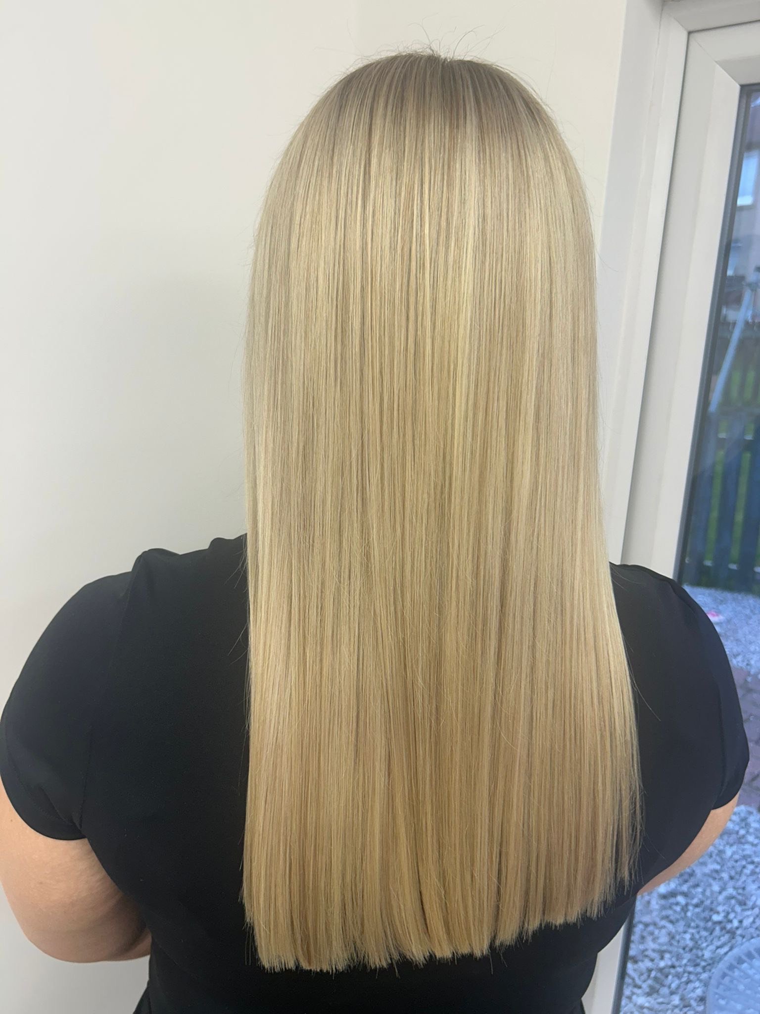 Back of a person with long, straight, blonde hair, wearing a black shirt, standing near a window.