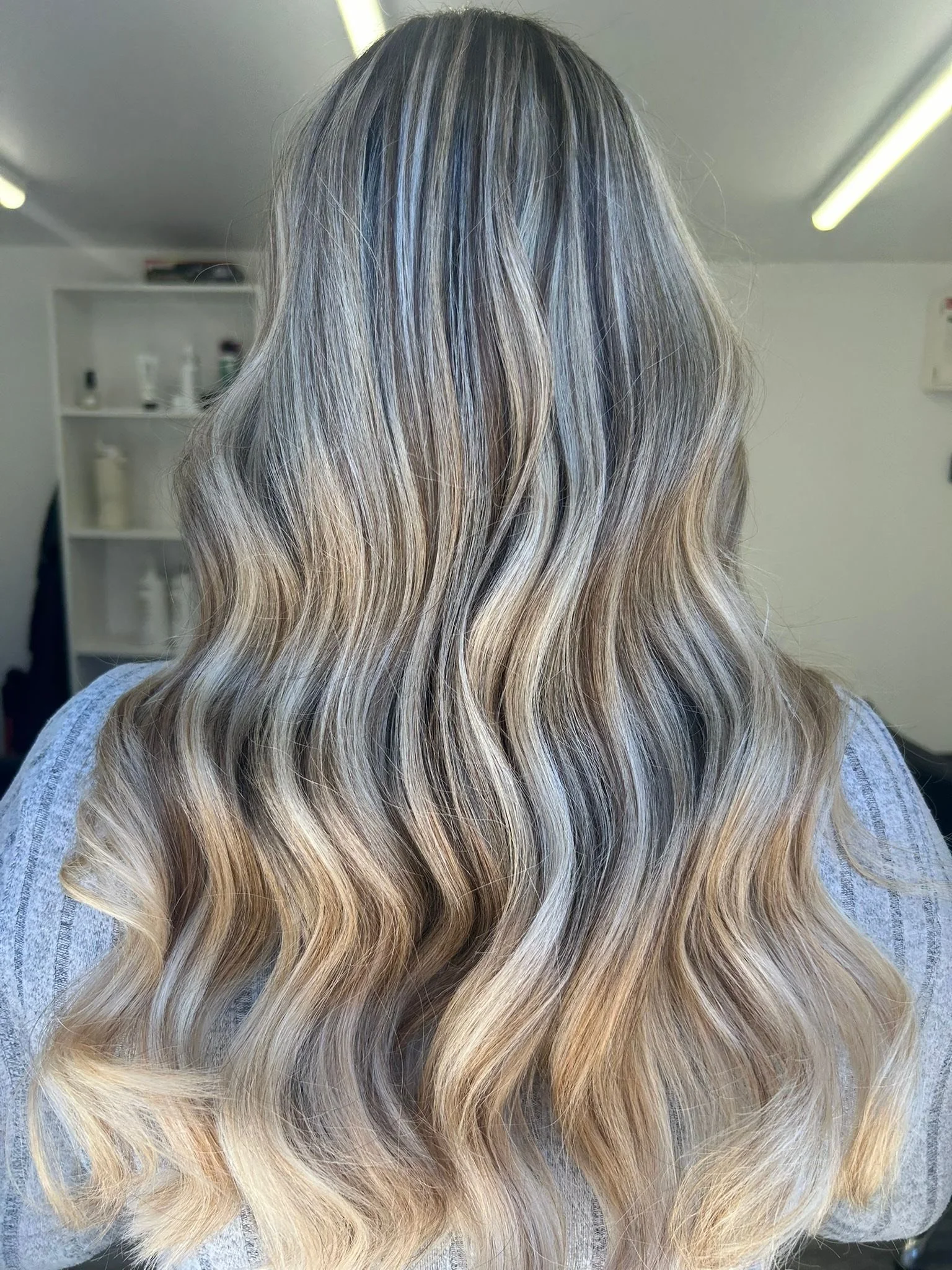 Back view of a woman with long, wavy, blonde hair with darker roots and lighter ends, in a salon or similar setting.