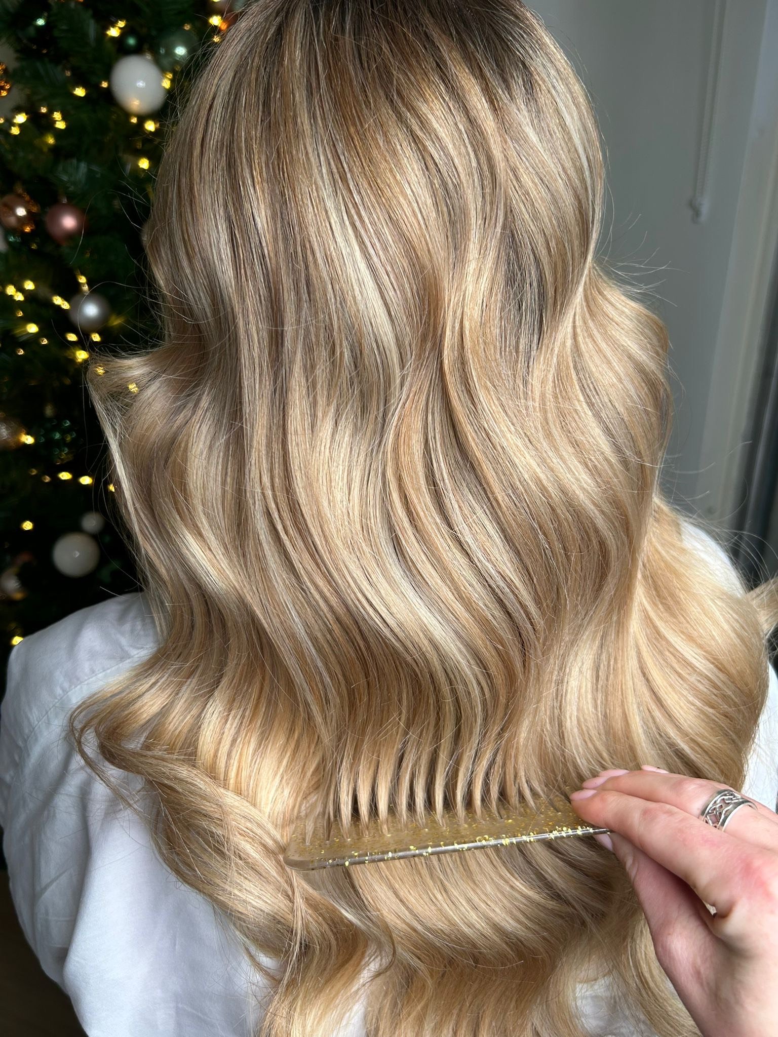 Close-up of a person with wavy blonde hair being combed with a wide-tooth comb, with a Christmas tree decorated with lights and ornaments in the background.