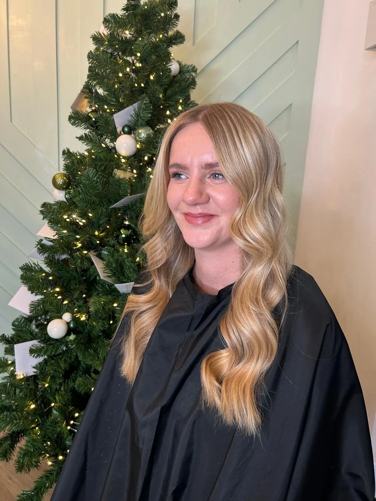 A woman with long, wavy blonde hair wearing a black salon cape, standing in front of a decorated Christmas tree with lights and ornaments.