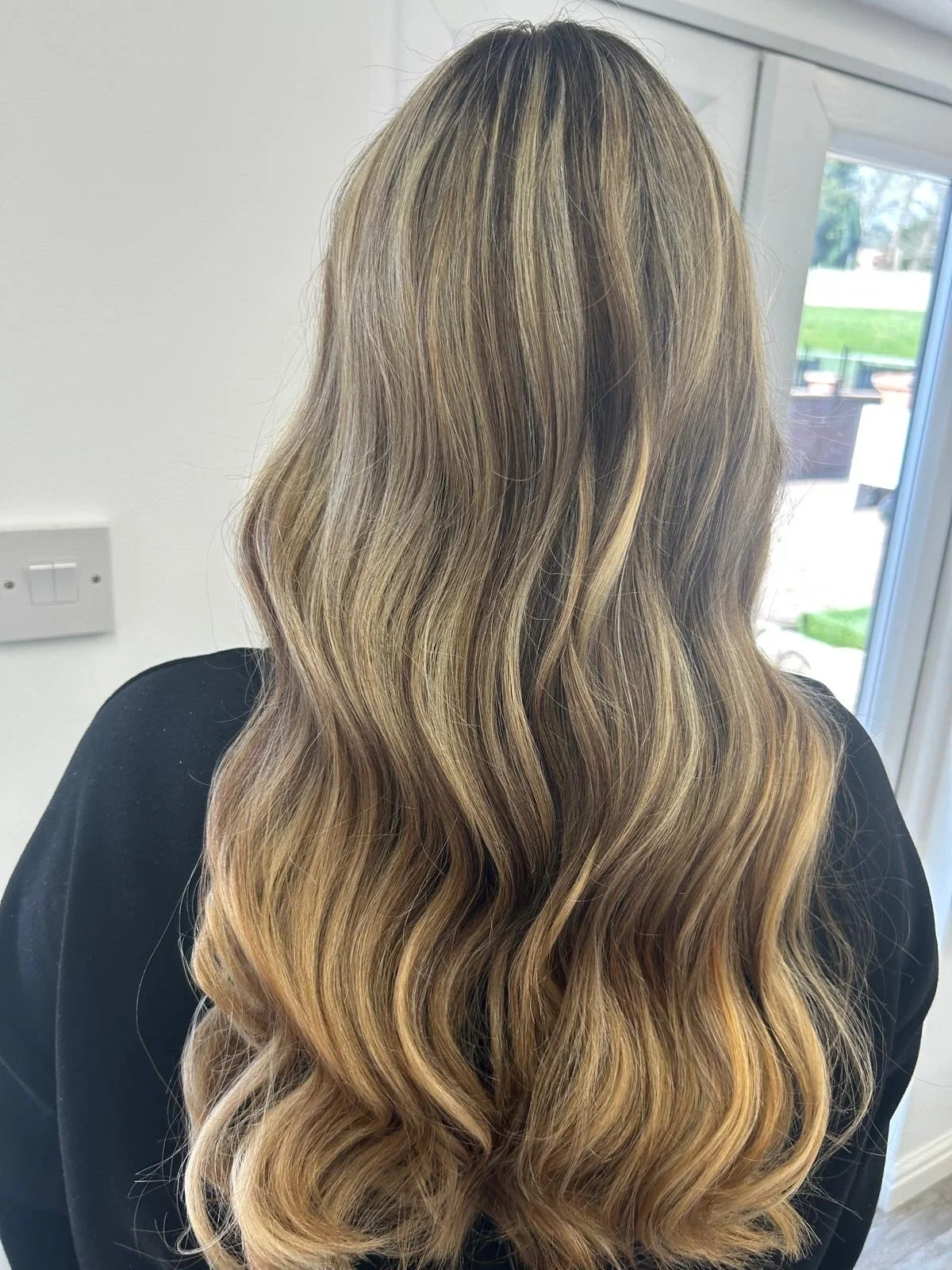 Back of a woman with long, wavy blonde hair with subtle highlights, wearing a black top, near a window with a view of a garden.