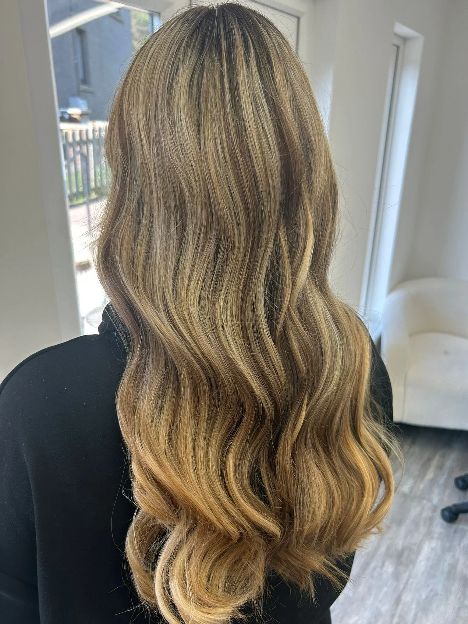 Back view of a woman with long, wavy, blonde hair standing inside near a window.