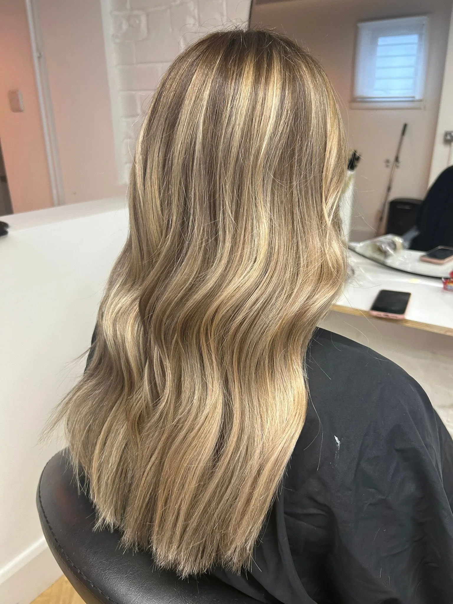 Back view of a woman with long, wavy blonde hair sitting in a salon chair.