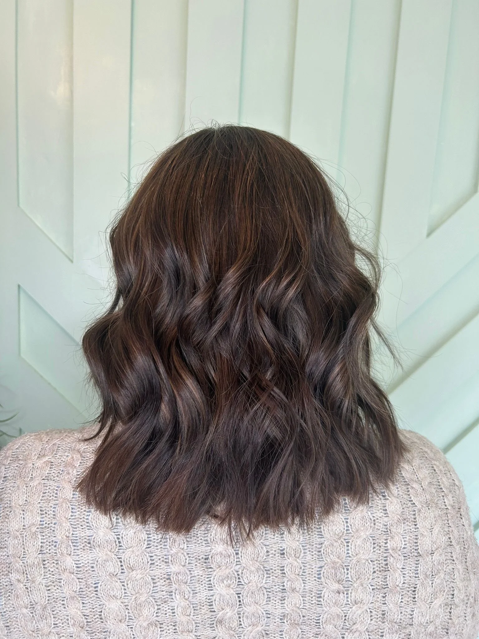 Back view of a person with medium-length wavy brown hair, wearing a light-colored knitted sweater, standing against a pale blue wooden background.