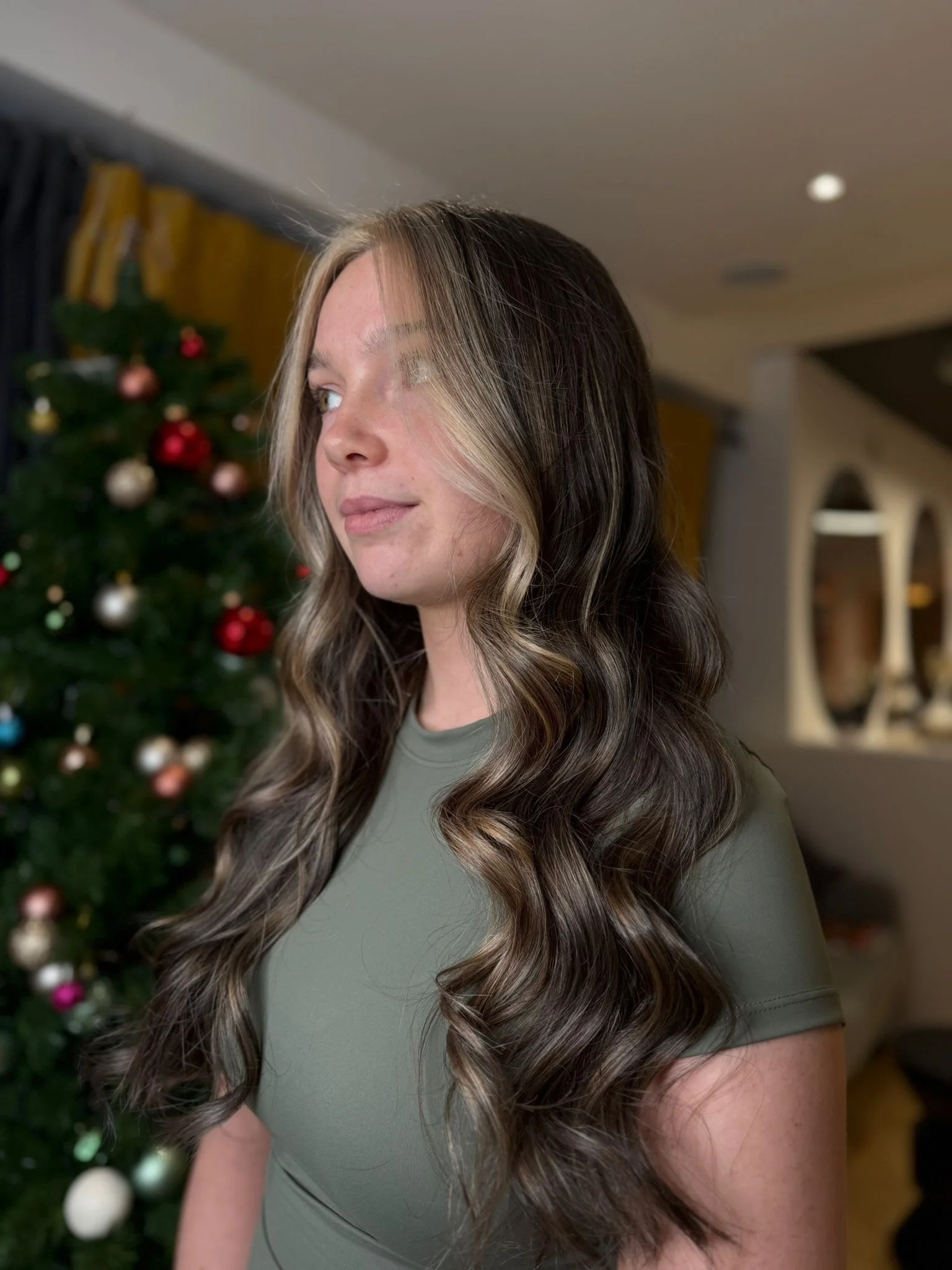 A woman with long, wavy brown hair with blonde highlights, wearing an olive-green top, standing in front of a decorated Christmas tree.