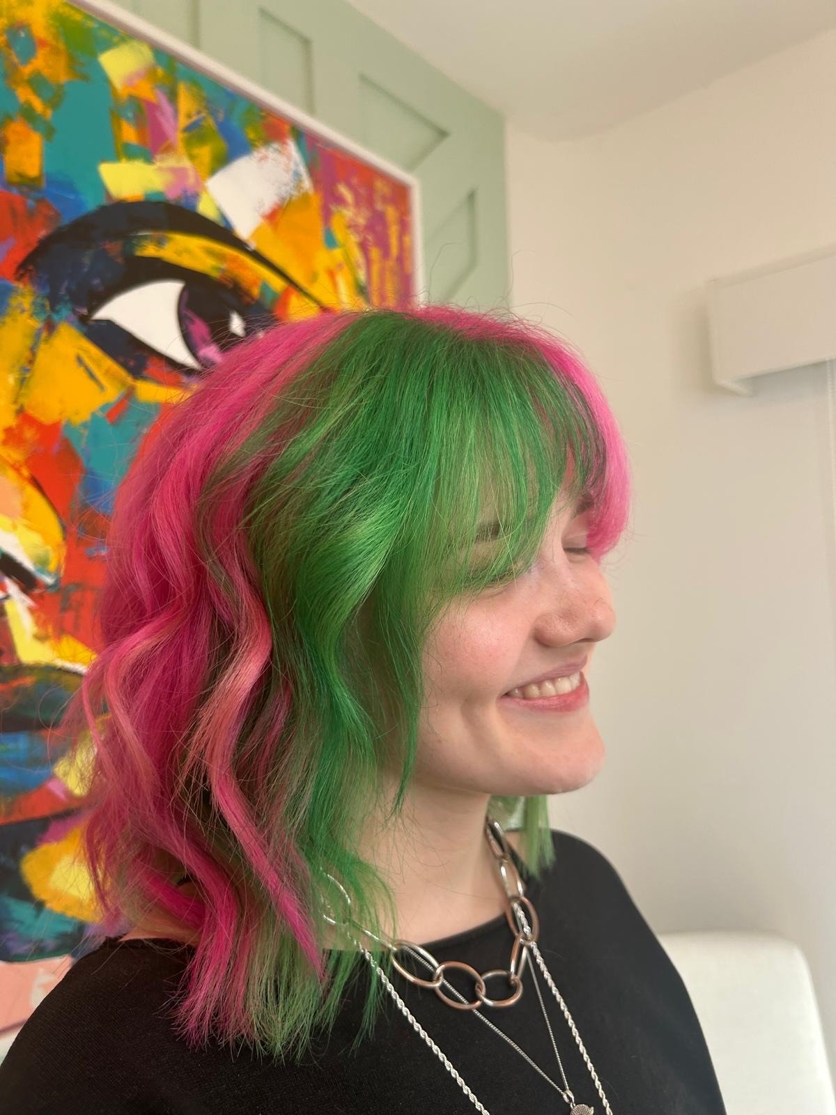A woman with shoulder-length hair dyed pink and green, smiling with her eyes closed, wearing a black top and silver jewelry, standing in front of a colorful abstract painting.