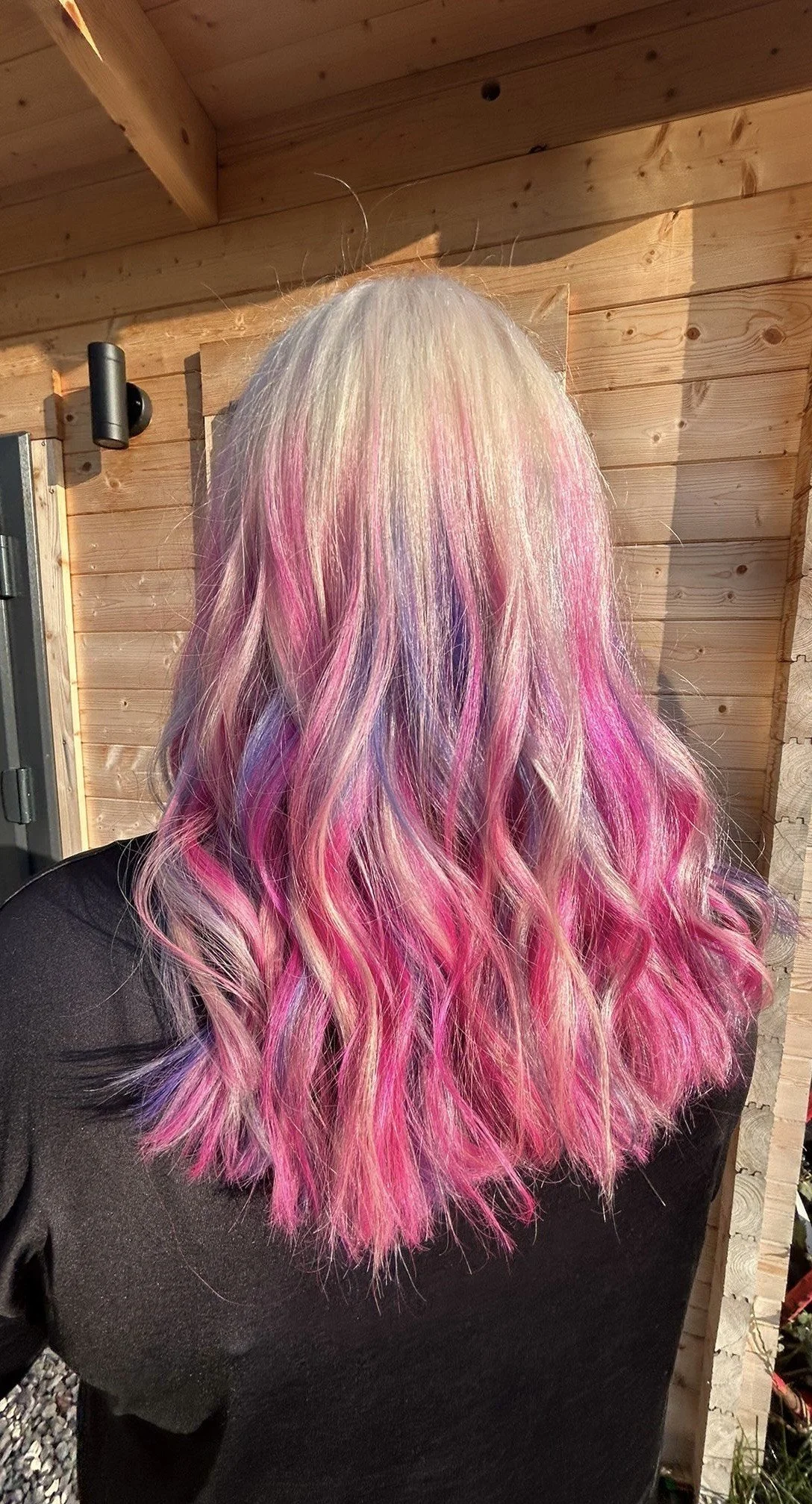Back of a person with long, wavy hair that is blonde at the top and transitions into pink and purple towards the ends, standing outdoors near a wooden wall.