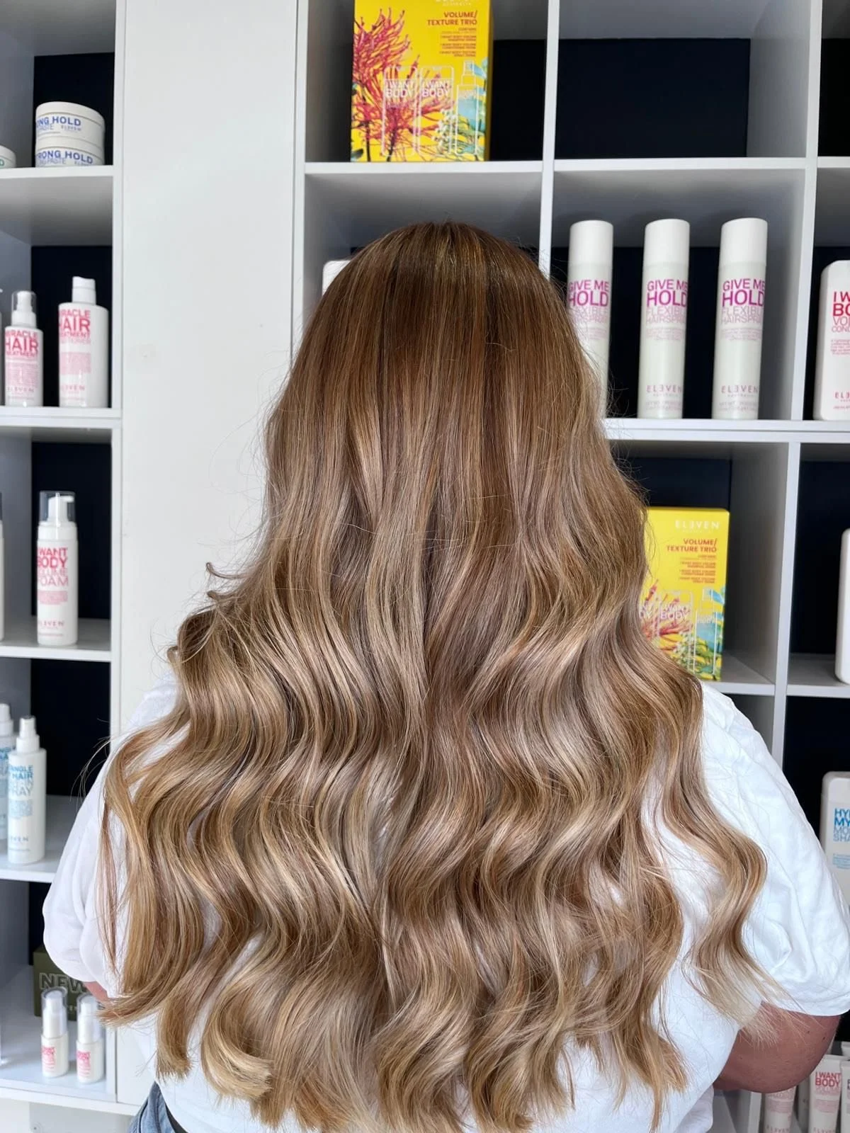 Back of a person with long, wavy, light brown hair standing in front of shelves displaying hair care products.