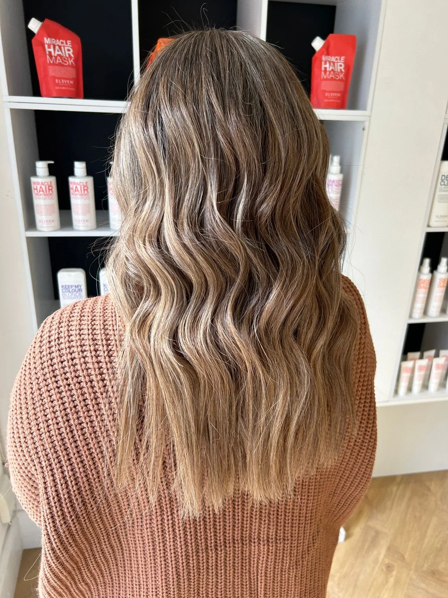 Back view of a woman with wavy, blonde hair wearing an orange sweater in a salon or store with hair care products on shelves.