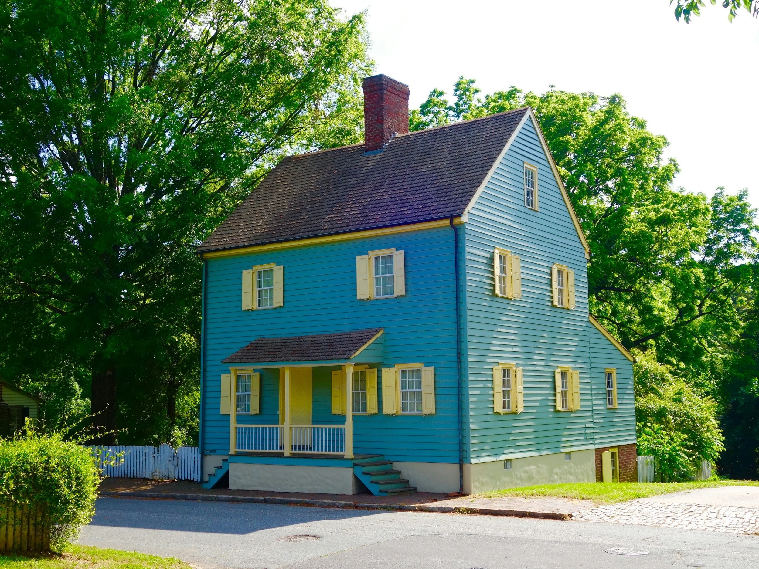A two-story house painted light blue with yellow window shutters, a small front porch, and surrounded by lush green trees.