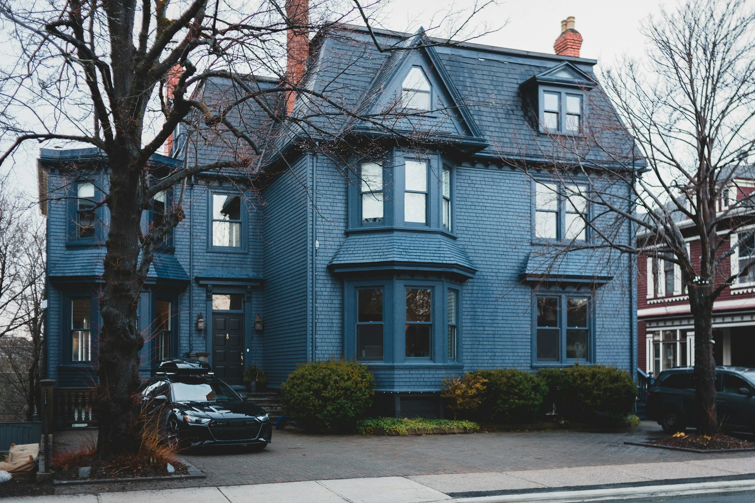 A blue, multi-story house with a dark roof, bay windows, trees, and parked cars in front.