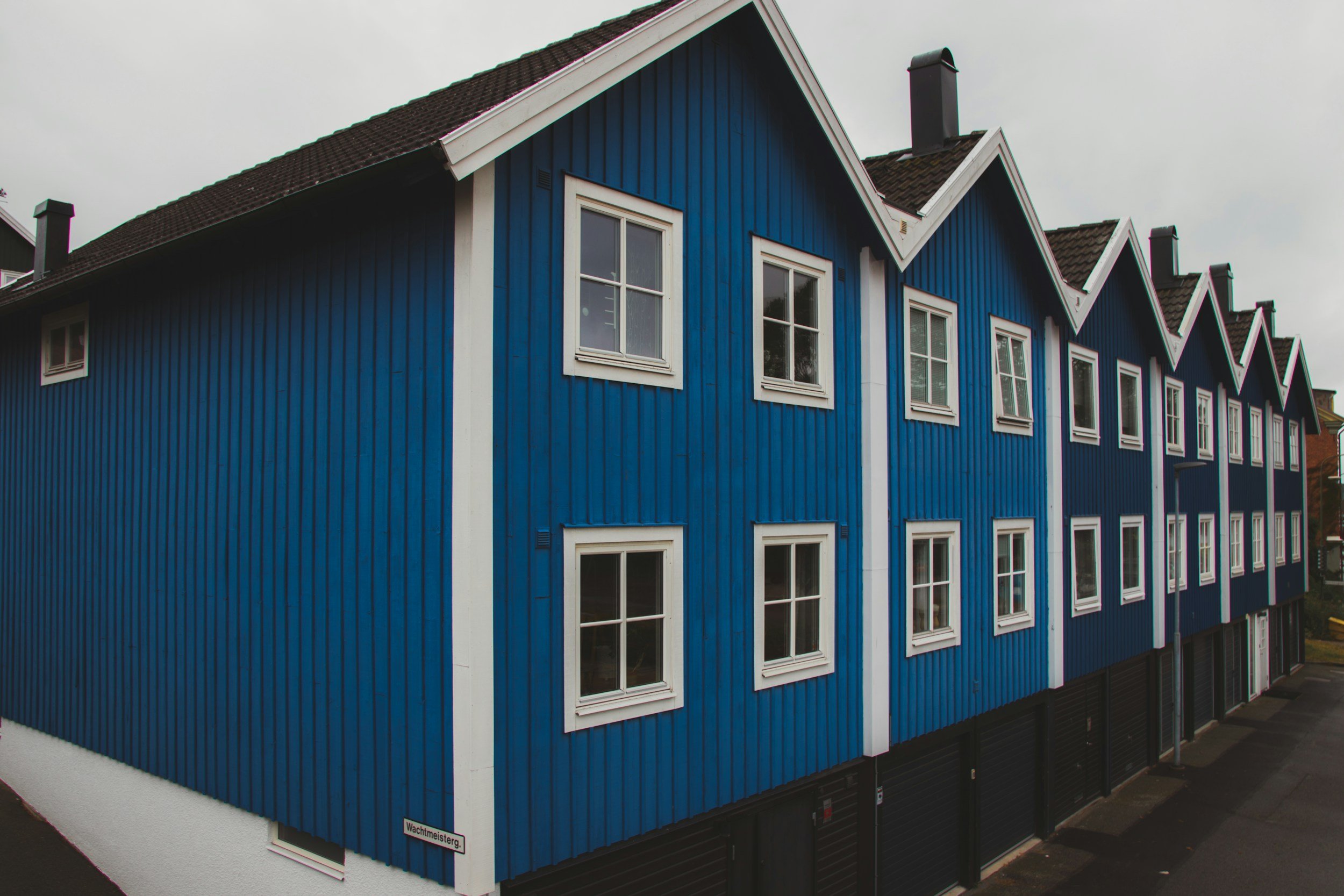 A modern, multi-story residential building with bright blue wooden exterior siding, white-framed windows, and a dark shingled roof, located along a street under an overcast sky.