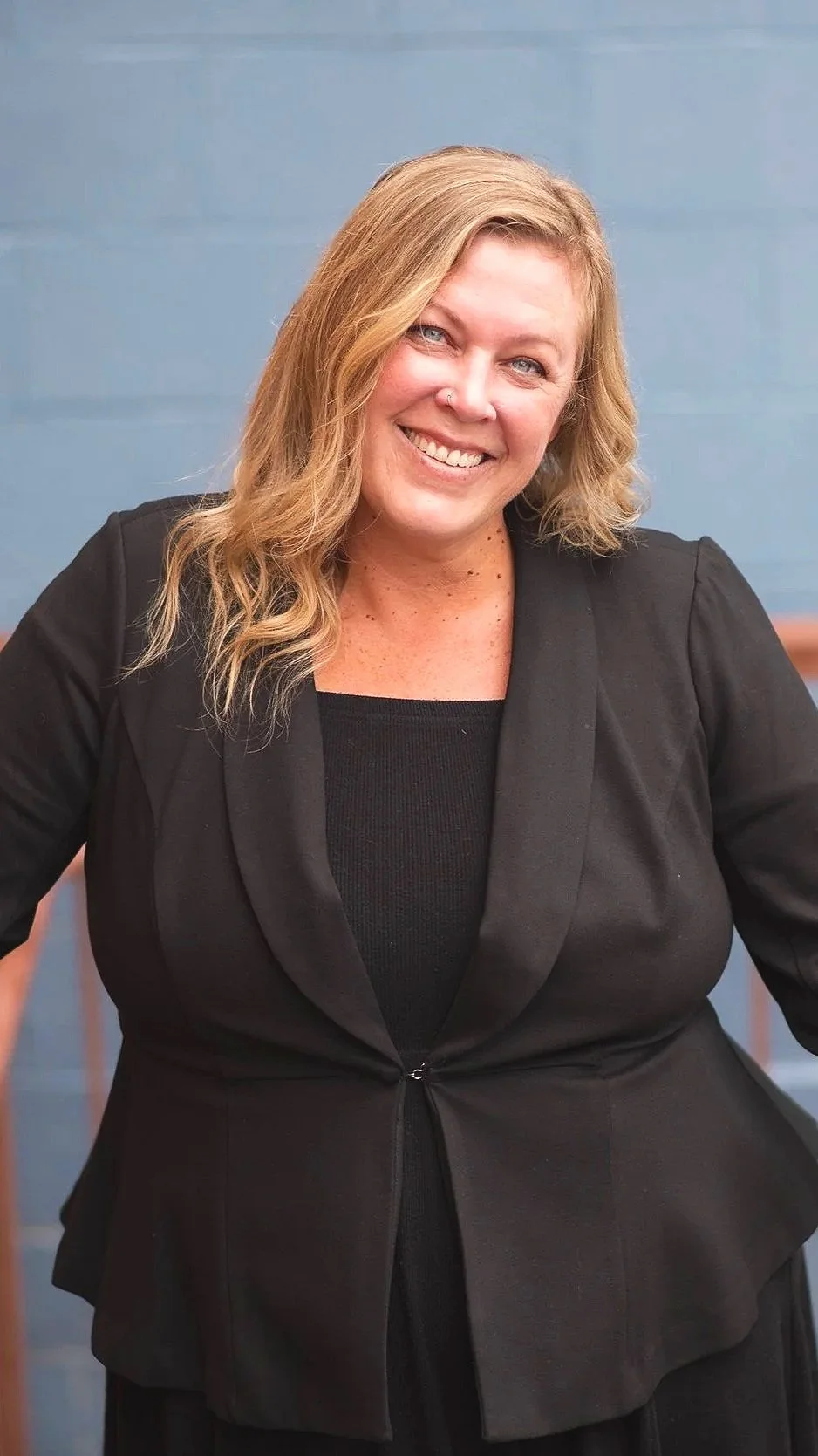A woman with wavy blond hair, wearing a black blazer and smiling, standing in front of a blue wall.