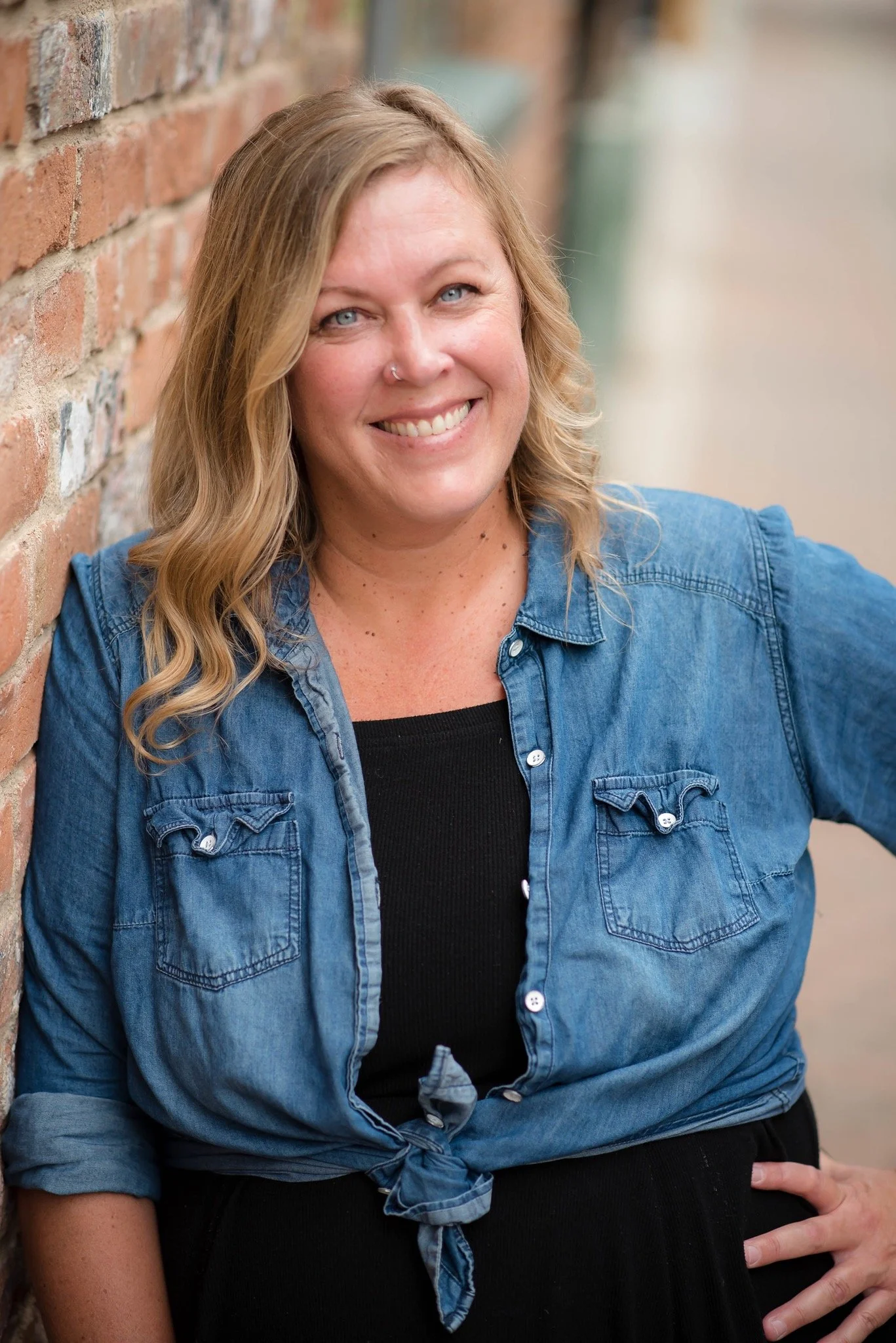 A smiling woman with wavy blonde hair wearing a tied blue denim shirt over a black top, leaning against a brick wall.